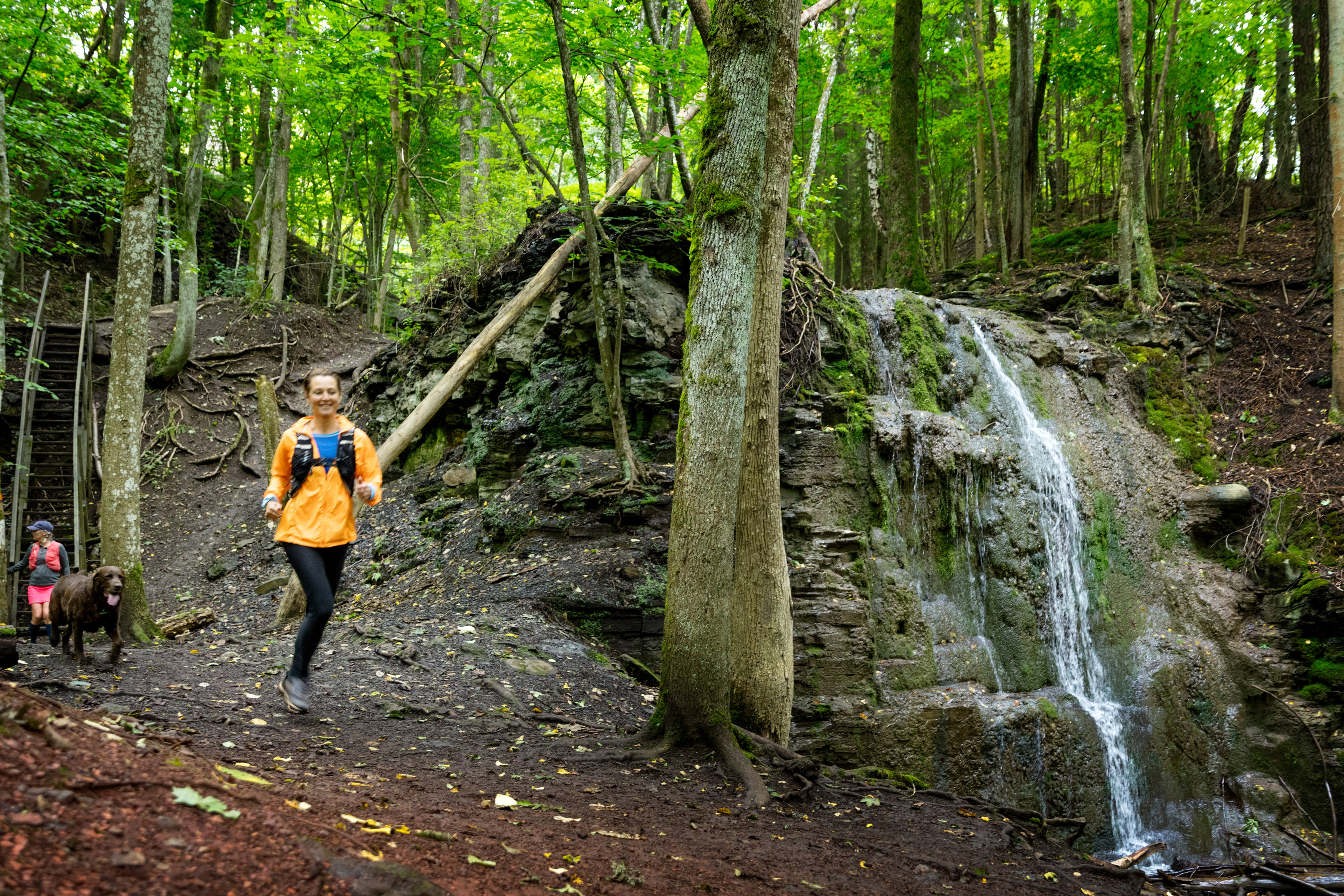 Kvinna springer intill vattenfallet Silverfallet bakom henne springer en brun hund. 