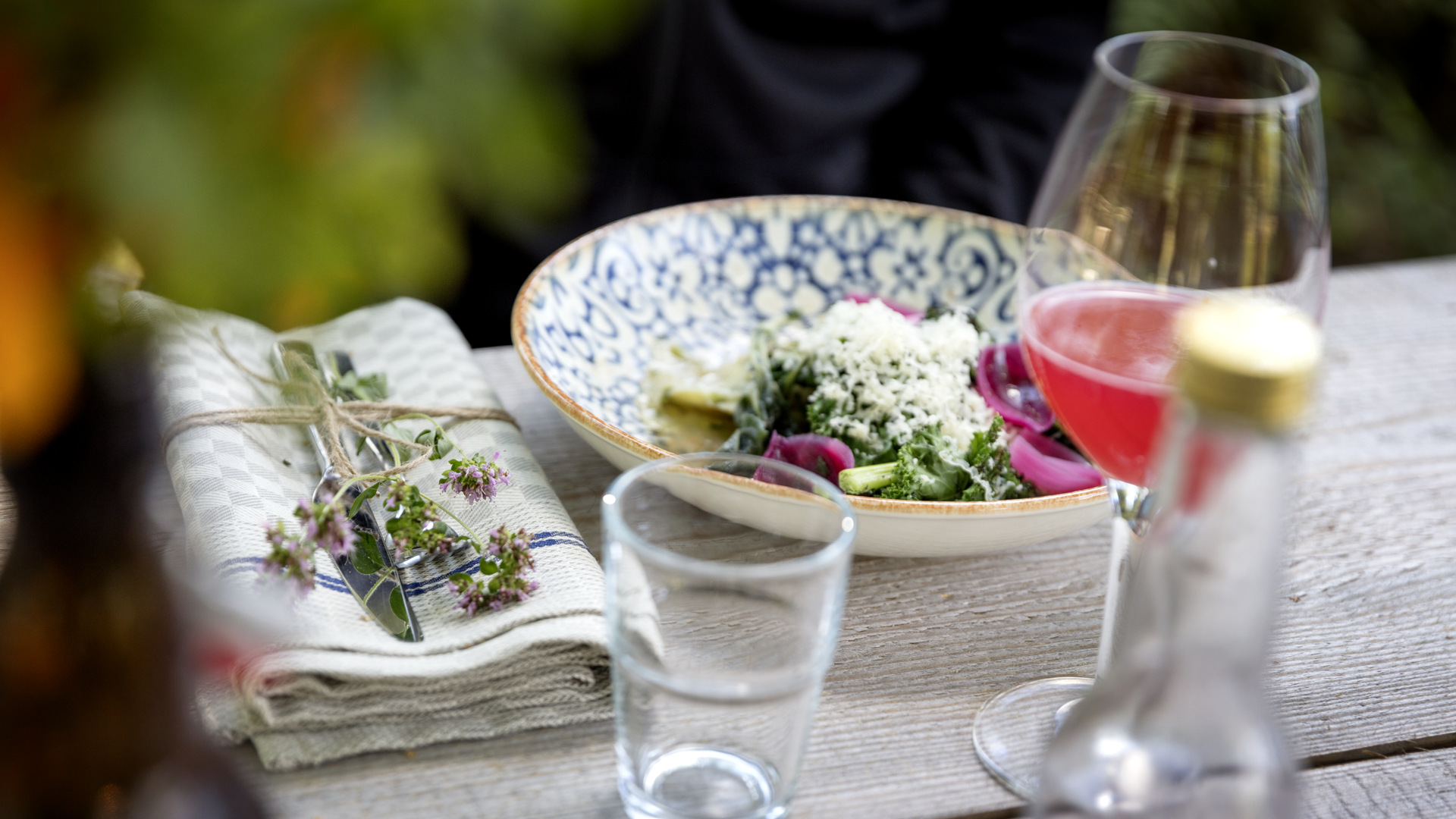 En vacker blåmönstrad tallrik med färgglad mat står framdukad på ett träbord med ett vinglas framför sig. Vinglaset innehåller en rosa och röd dryck. 