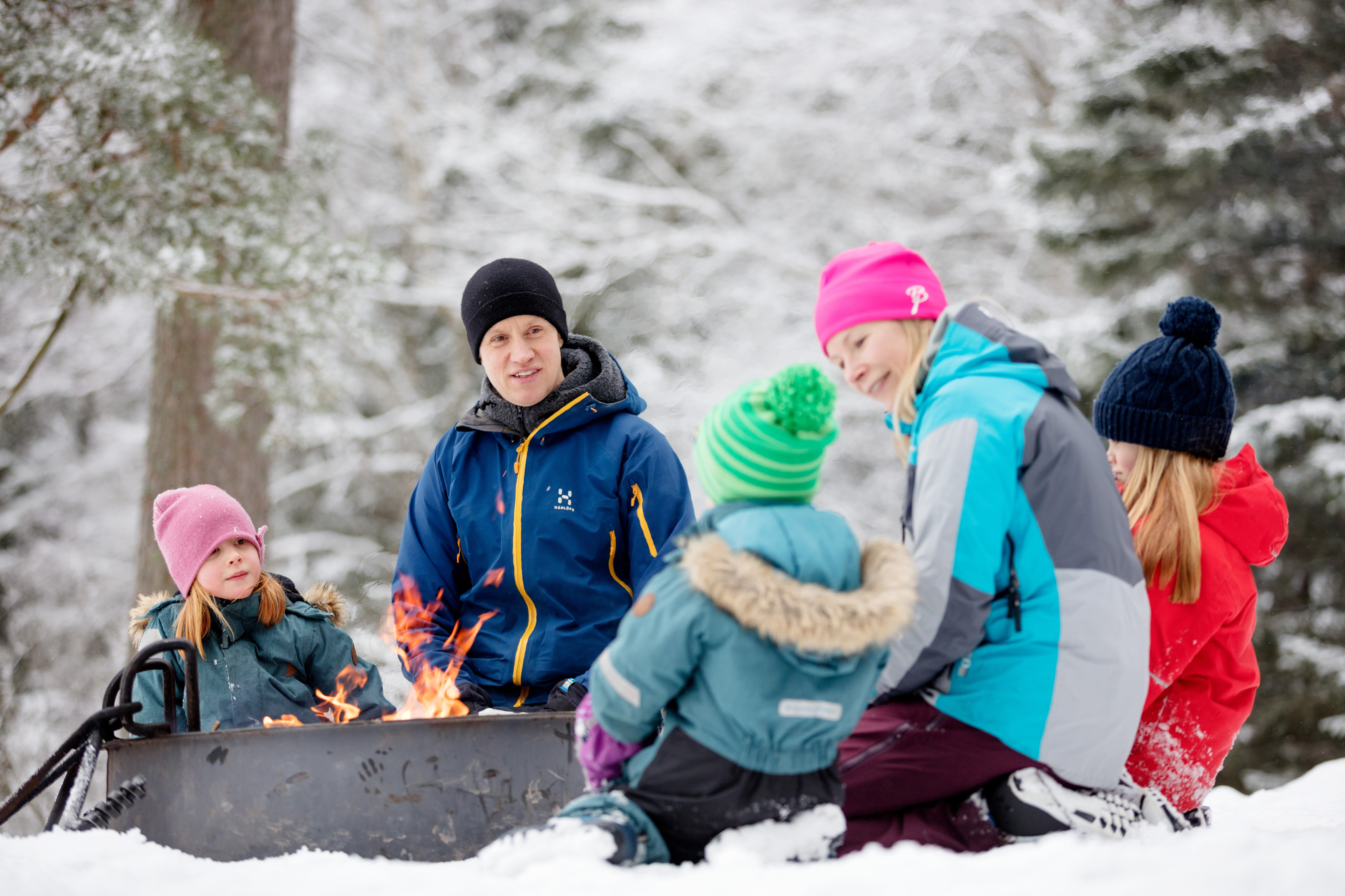 En familj på två vuxna och tre barn sitter vid en grillplats i snön och har gjort upp eld.