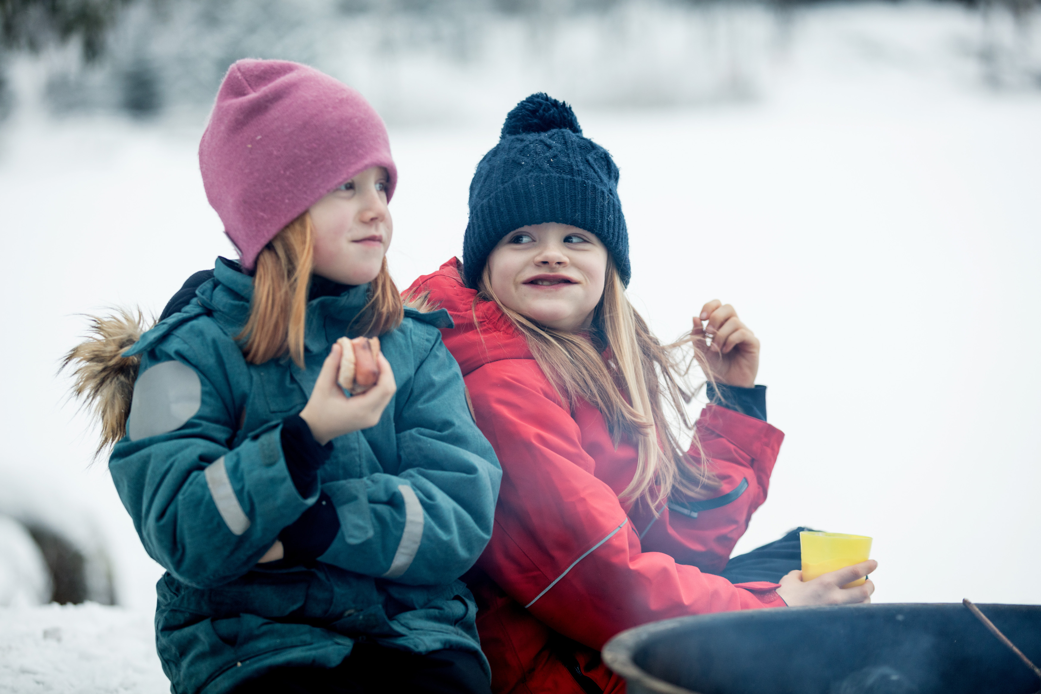 Family sitting by the fire surrounded by a winter landscape at Billingen moutain. Two girls are eating hot dogs. 