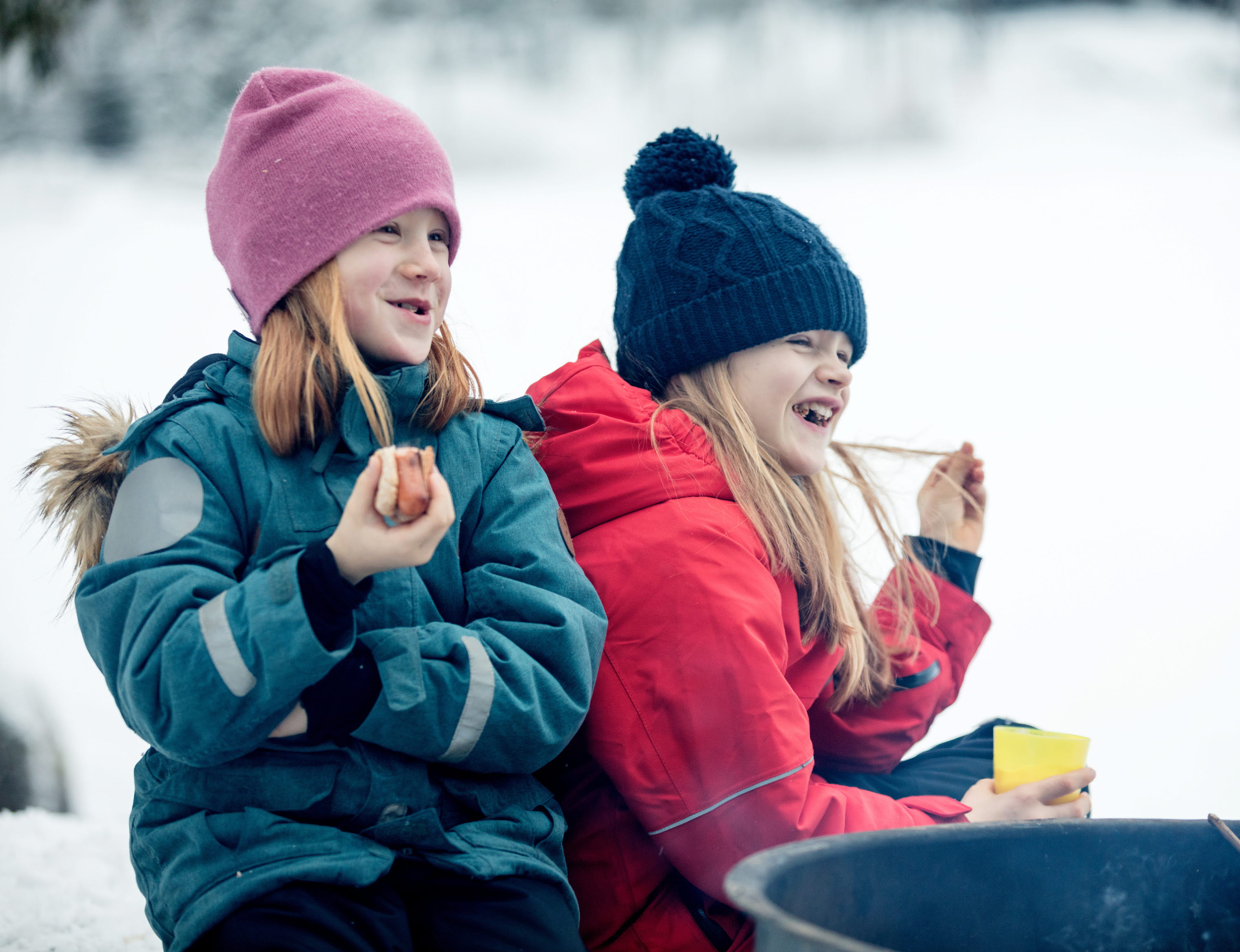 Family sitting by the fire surrounded by a winter landscape at Billingen moutain. Two girls are eating hot dogs and laughing.