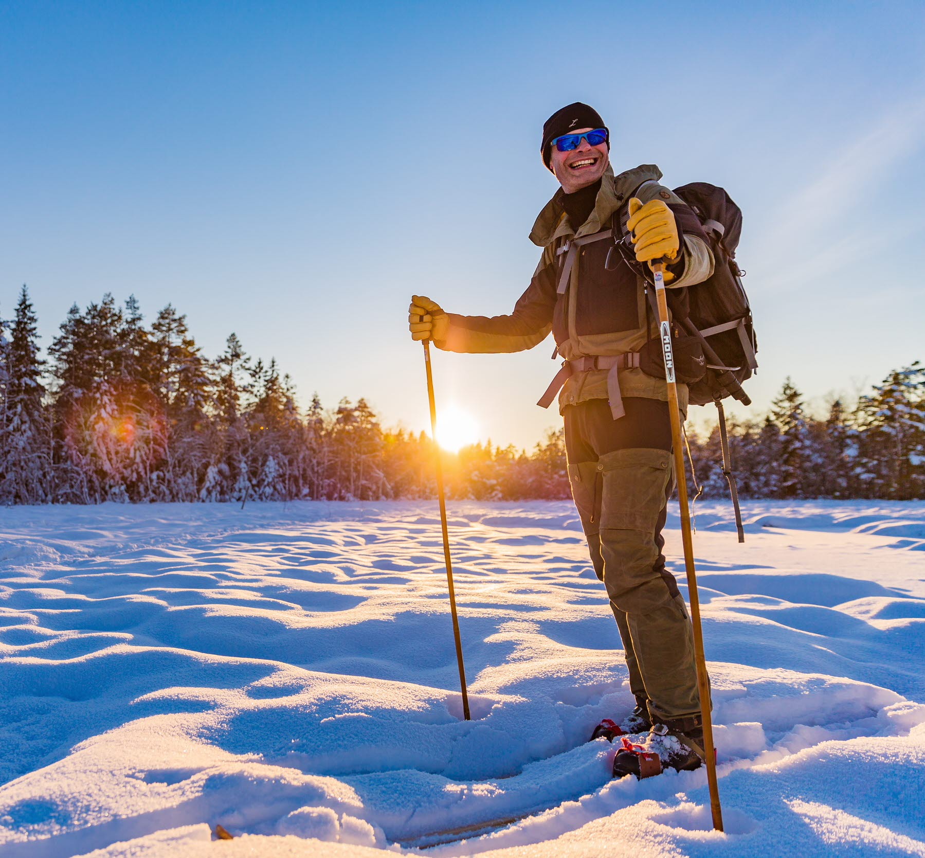 Soligt vinterlandskap där en person står i snön med skidor på fötterna och stavar i händerna. 