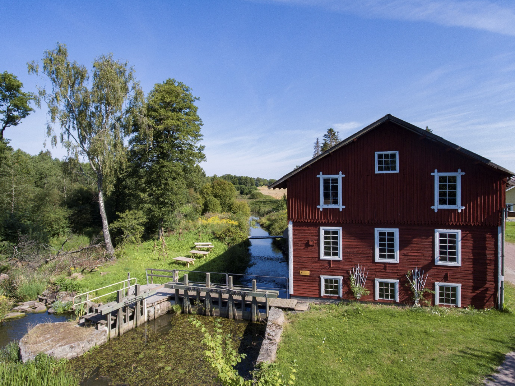 En gammal röd kvarnbyggnad med vita spröjs vid en å med gröna omgivningar. 