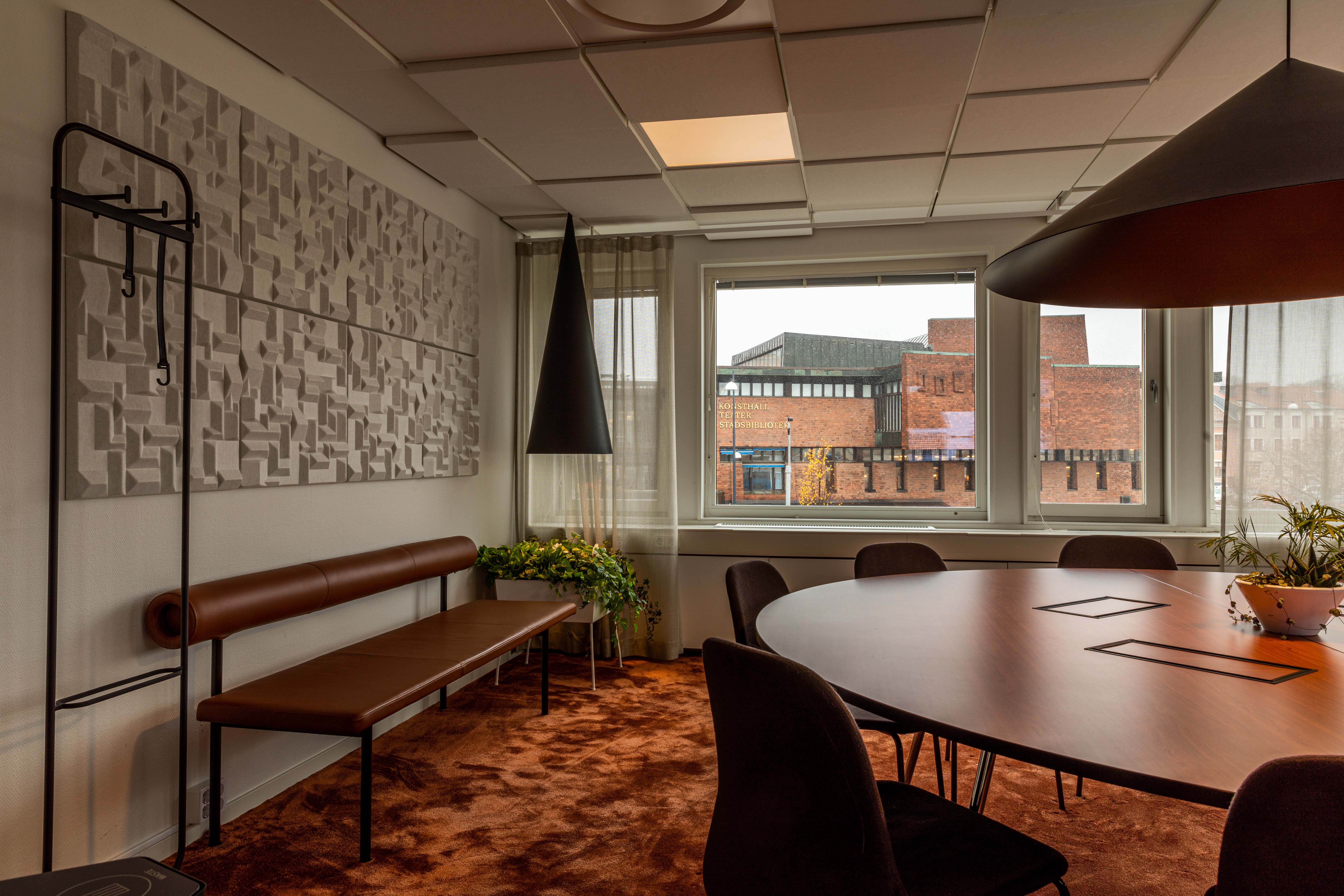 Ett konferensrum med ett rundt bord i förgrunden, en bänk och genom fönstret ser man Skövde kulturhus.