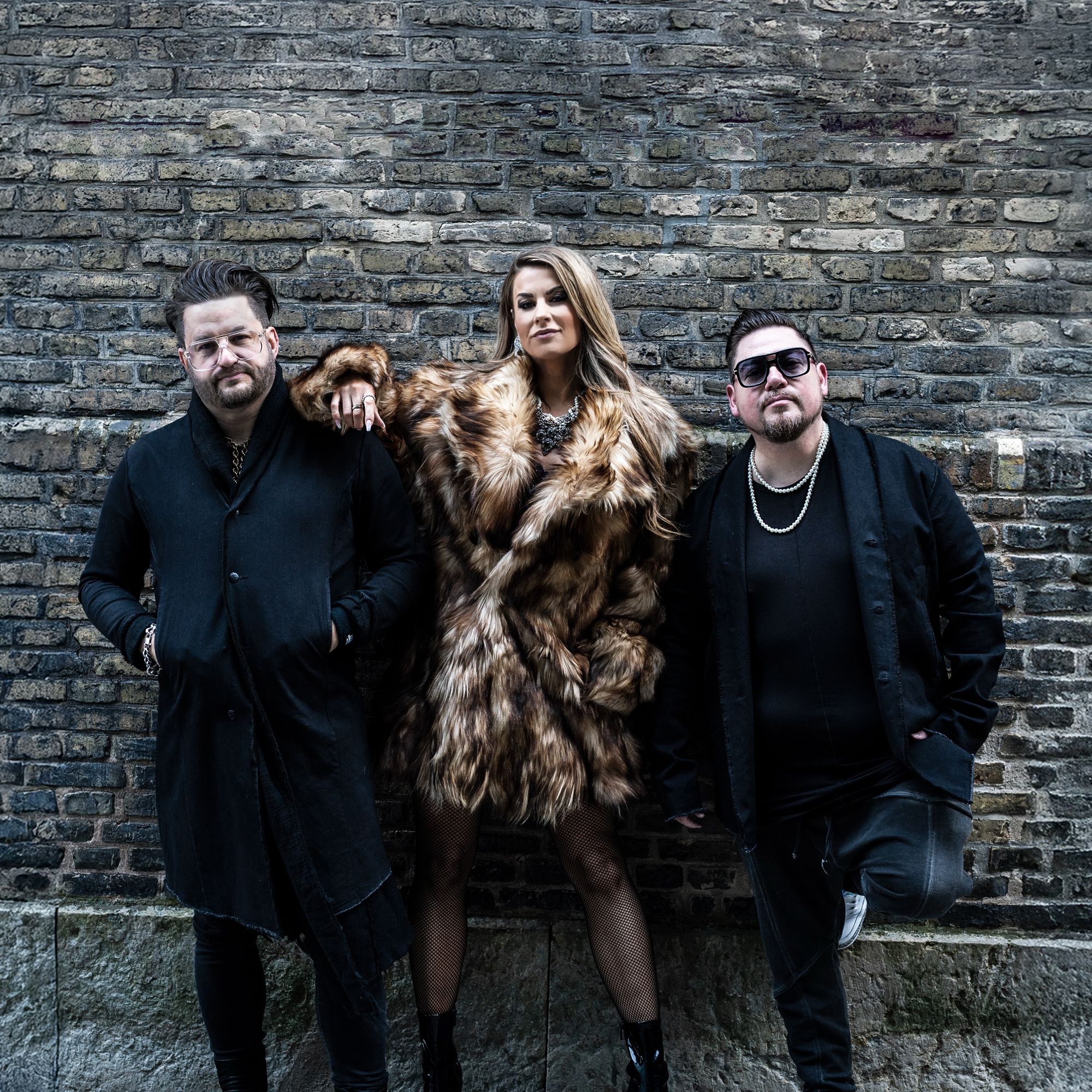 Ett band med en tjej och två killar framför en tegelvägg. 