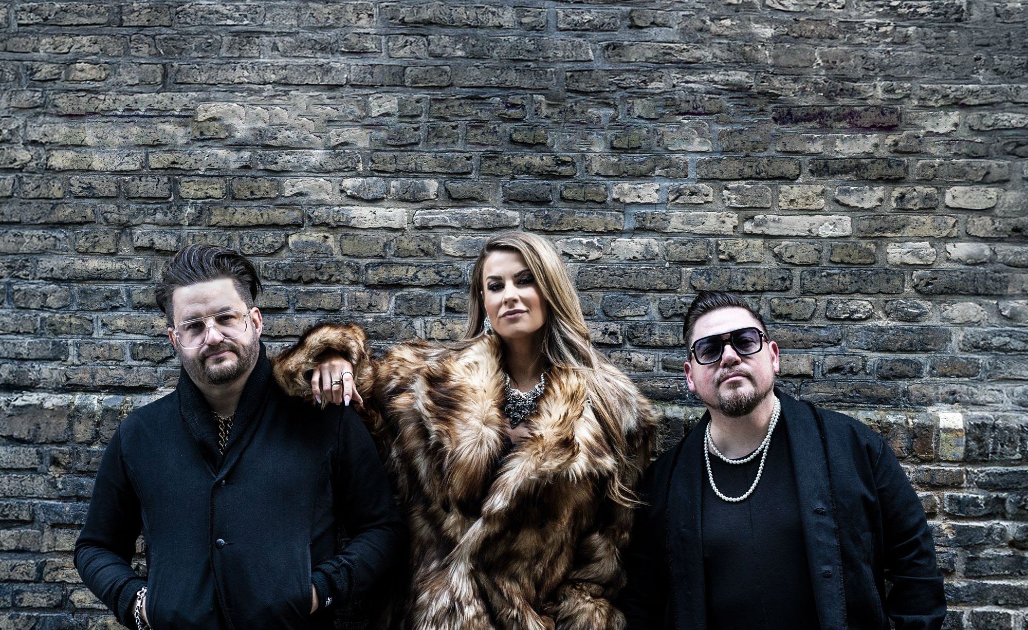 Ett band med en tjej och två killar framför en tegelvägg. 