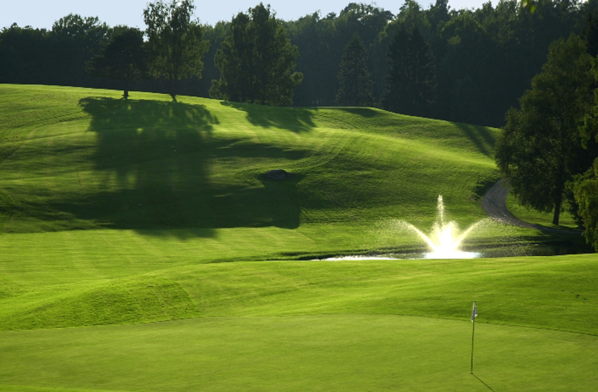 En vy över Billingens golfbana, grönt gräs och en damm med en fontän i syns till höger i bilden.