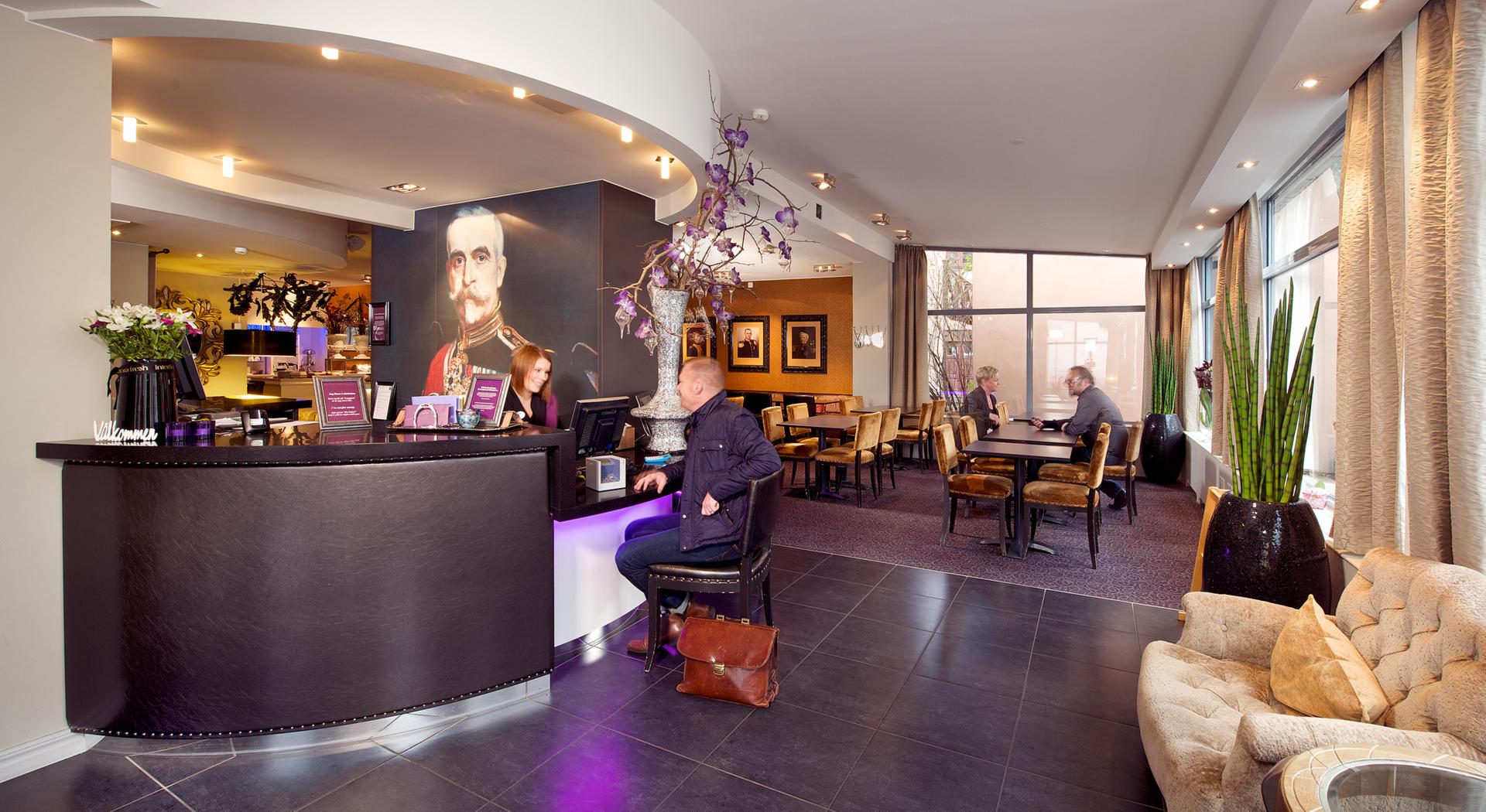 A man is sitting on a chair in the hotel reception. Gets help from staff to check in. In the background you can see part of the dining and breakfast room where two people are sitting and talking.