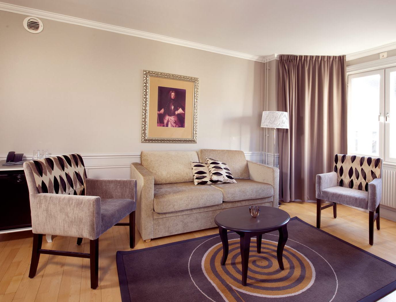 En soffa och två fåtöljer framför stort fönster. I nederkant syns en hotellrummets säng och ett sängbord.  