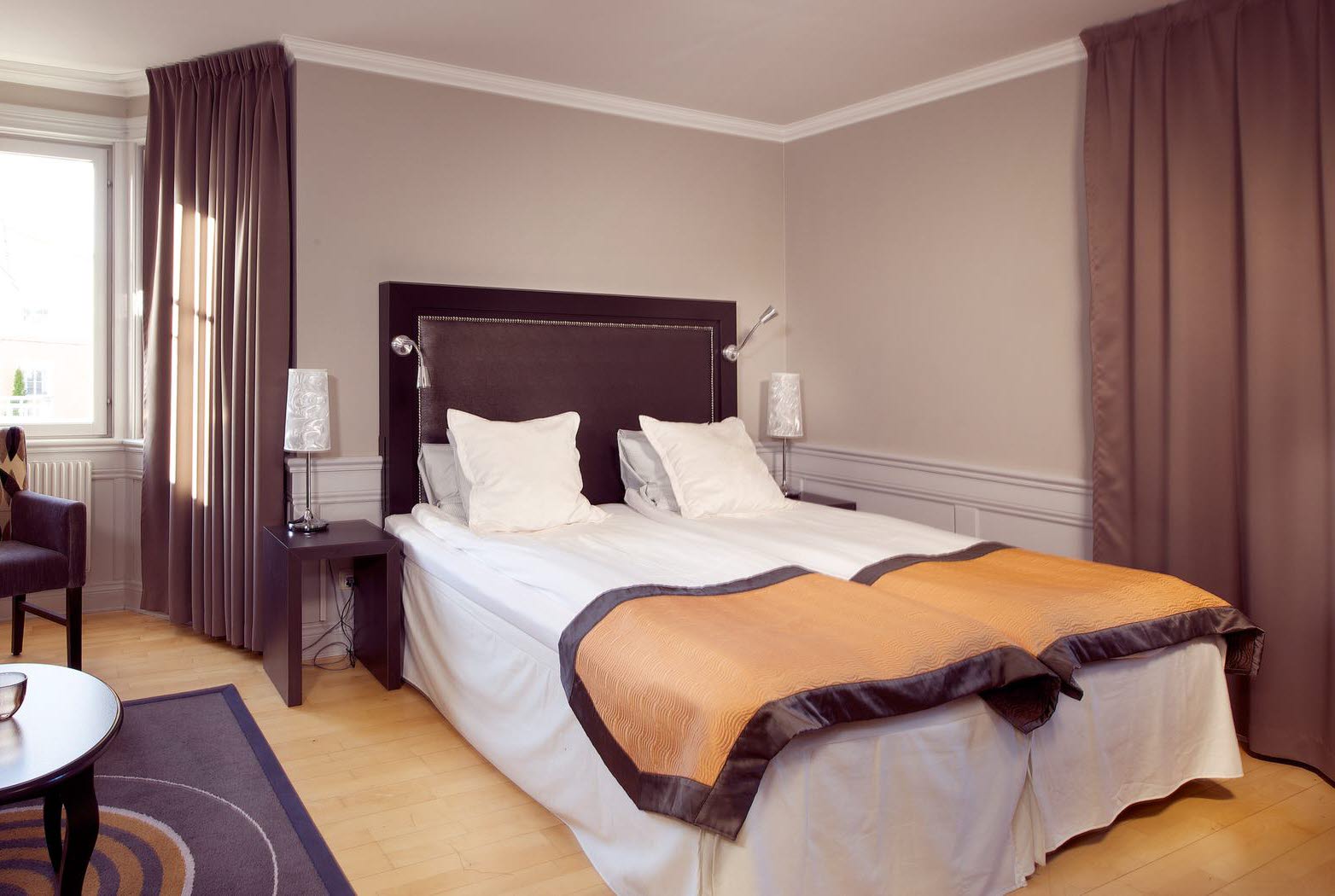 En vackert bäddat dubbelsäng med kuddar och filtar i ett stort hotellrum. Till vänster i bild syns en fåtölj framför ett stort fönster med gardinerna dragna åt sidorna.