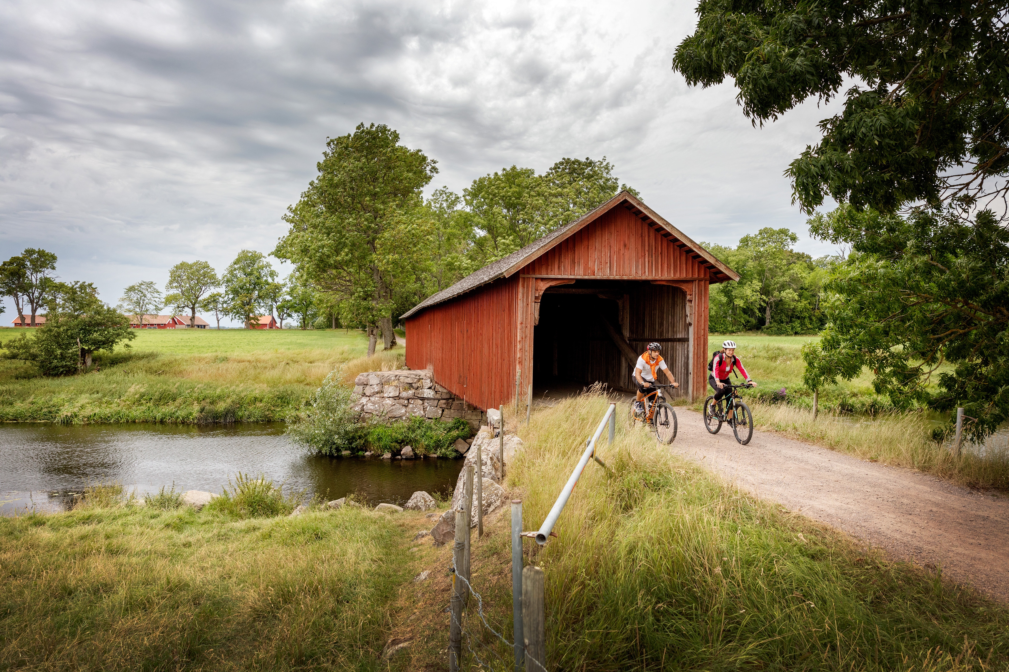 En man och en kvinna cyklar på en grusväg vid det unika långa brohuset, Vaholms brohus.
