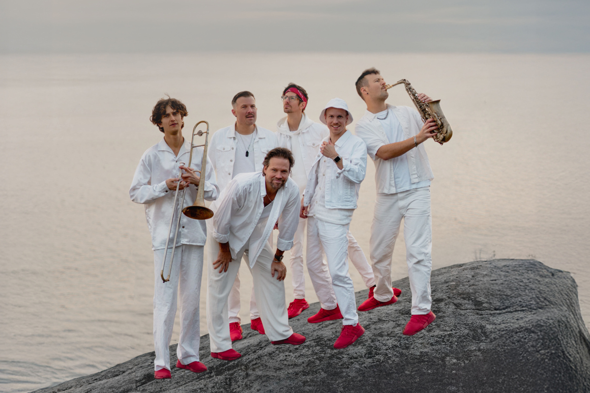 En grupp av människor med vita kläder och röda skor på en sten framför ett hav. Två av killarna håller i blåsinstrument. 