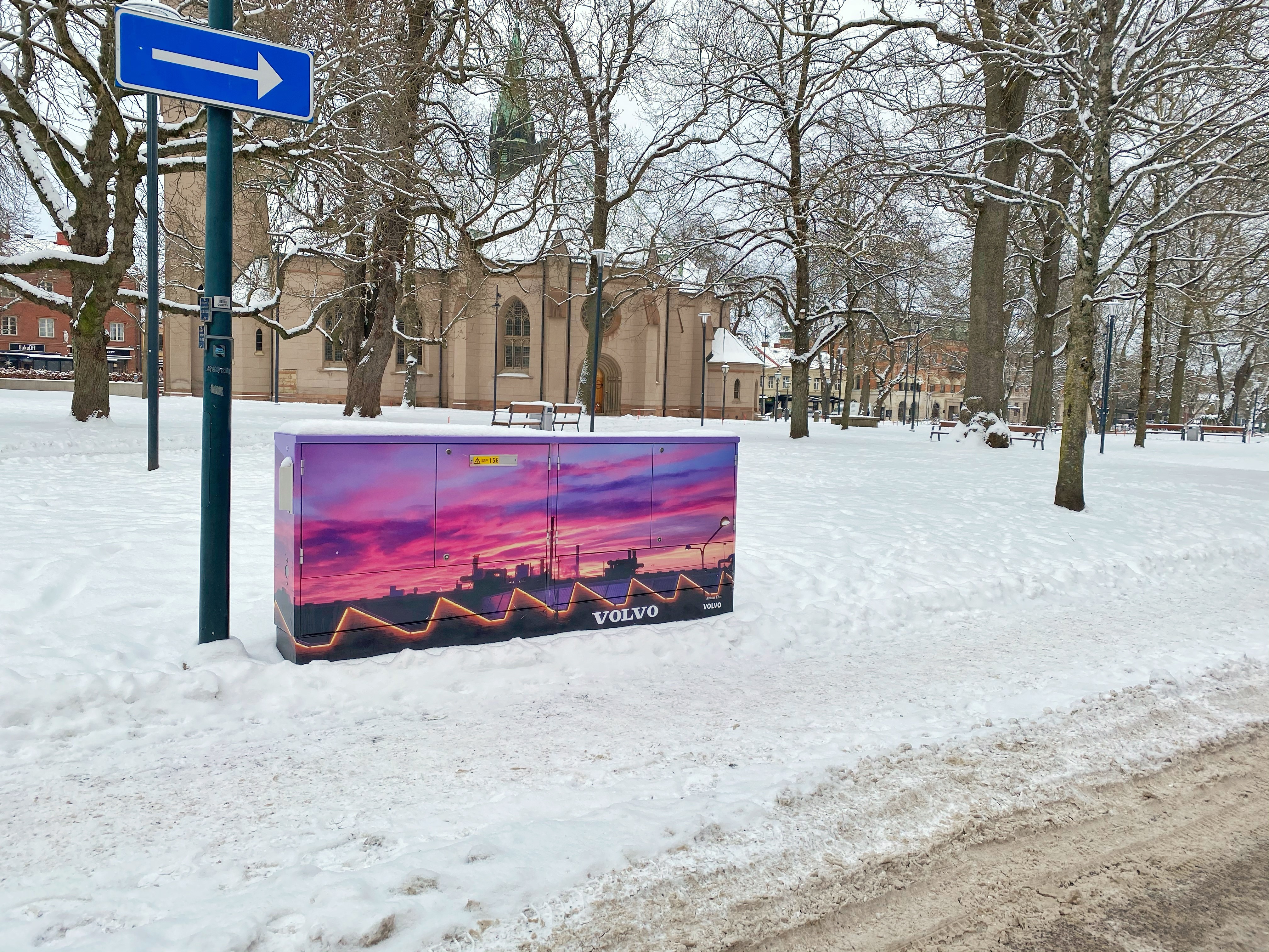En vintrig bild från kyrkparken med kyrkan i bakgrunden där ett elskåp som klätts med en bild på Volvofabriken i skymning syns.