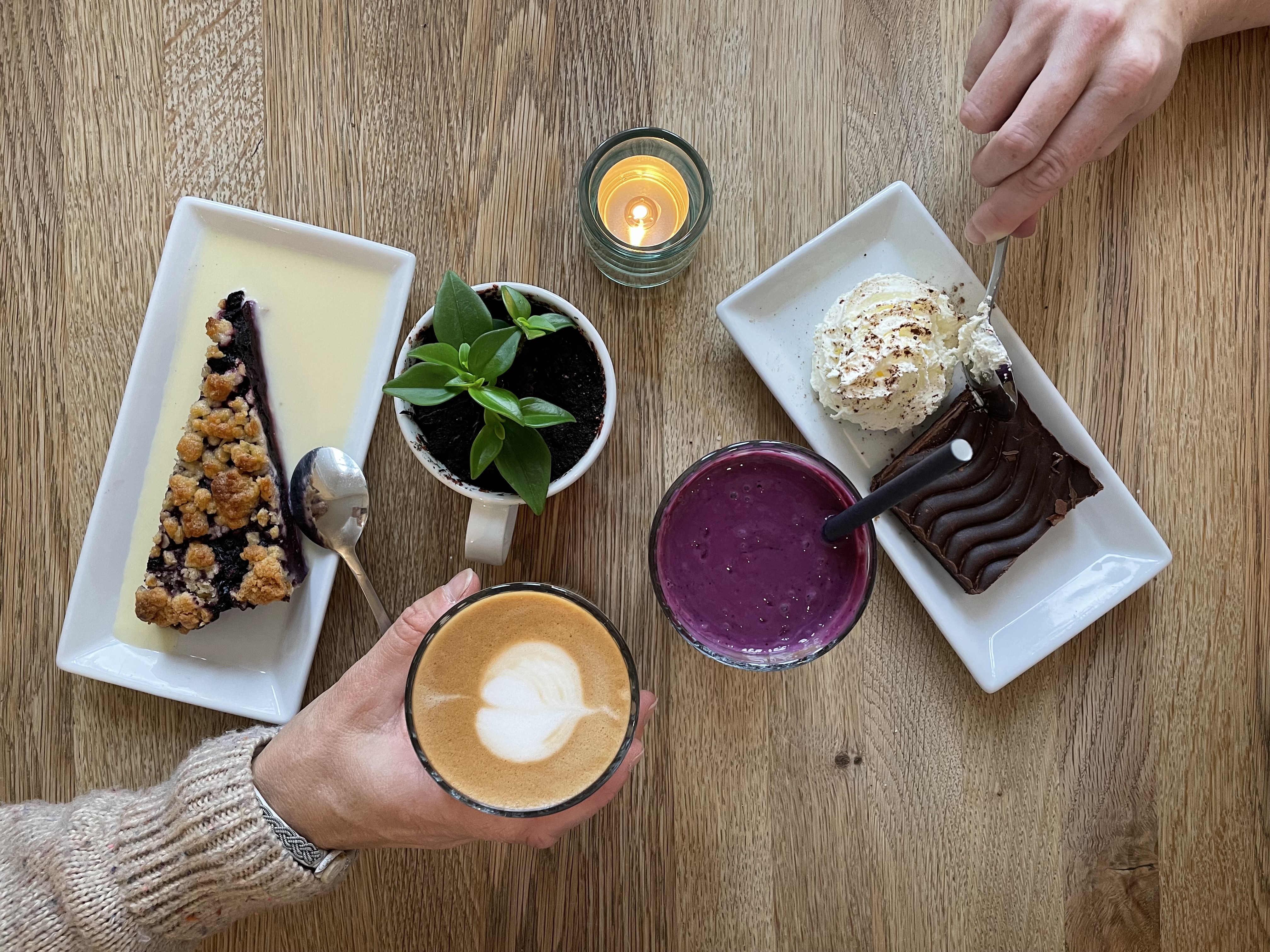 Ett bord med fat som det ligger kakor på. På bordet står också en kaffe latte och en smothie. Man kan se en hand som håller i glaset med kaffe latte och en hand som håller i en sked på väg att ta en bit av en kaka.