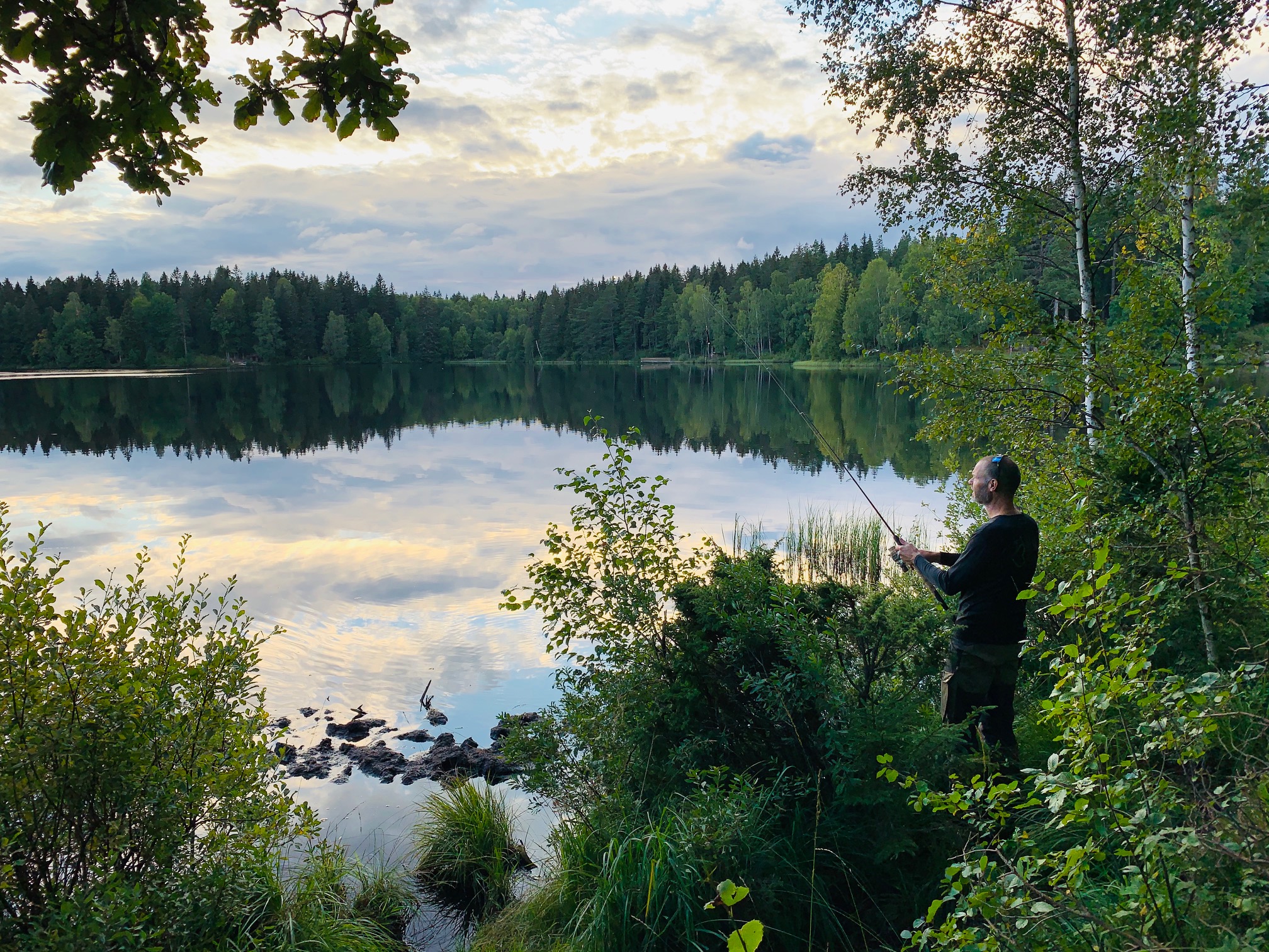 En man står vid kanten av en spegelblank sjö och fiskar i skymningen. 