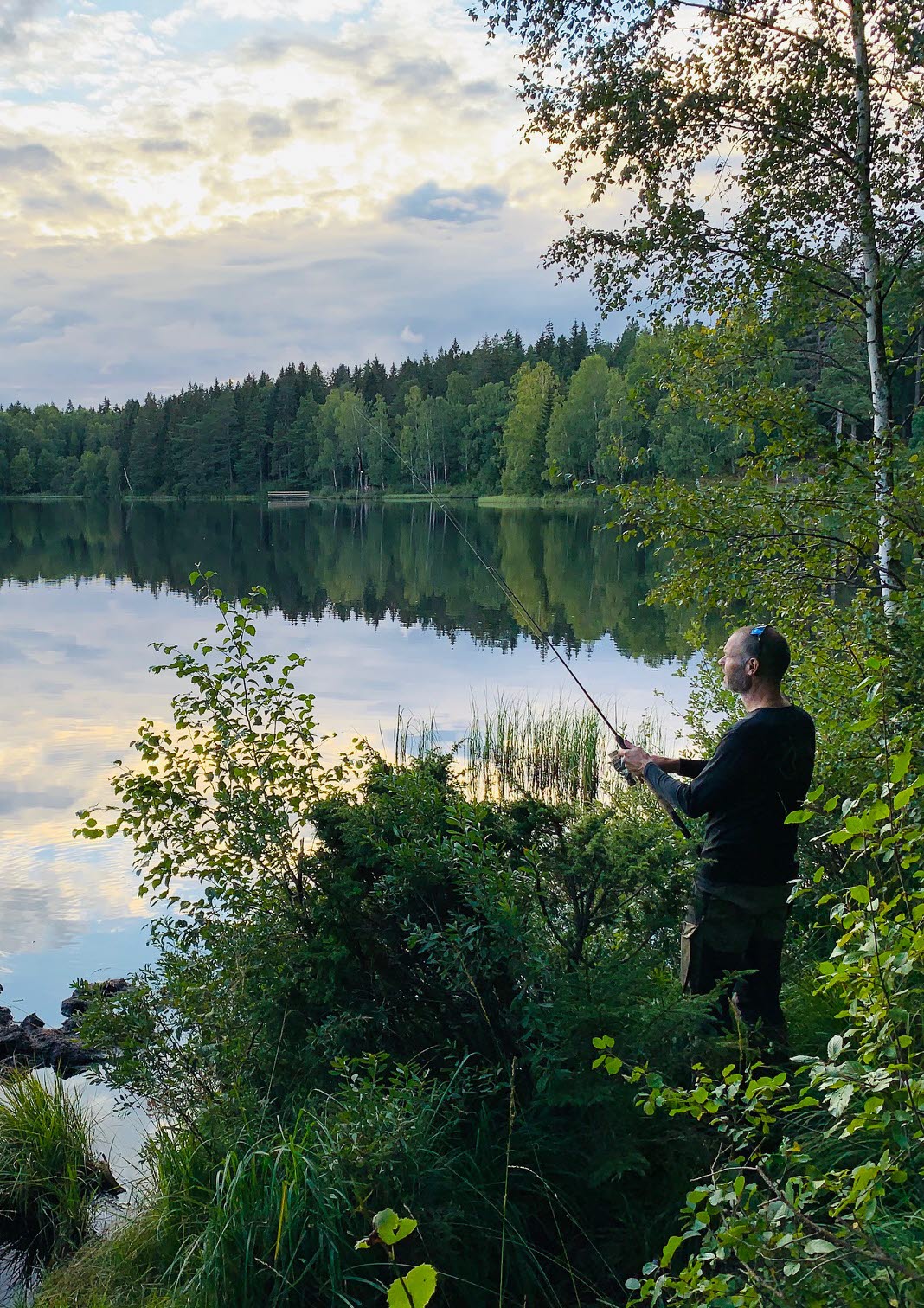 En man står vid kanten av en spegelblank sjö och fiskar i skymningen. 