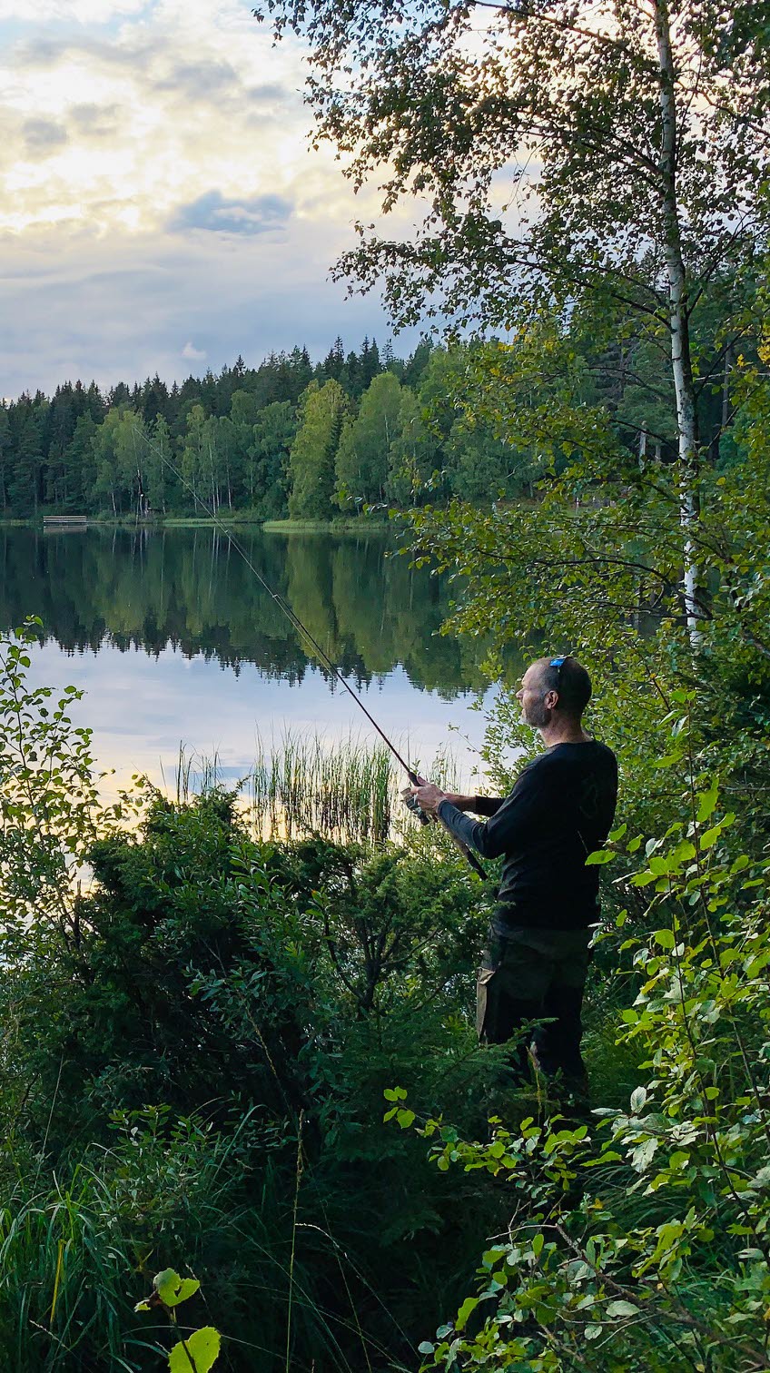 En man står vid kanten av en spegelblank sjö och fiskar i skymningen. 