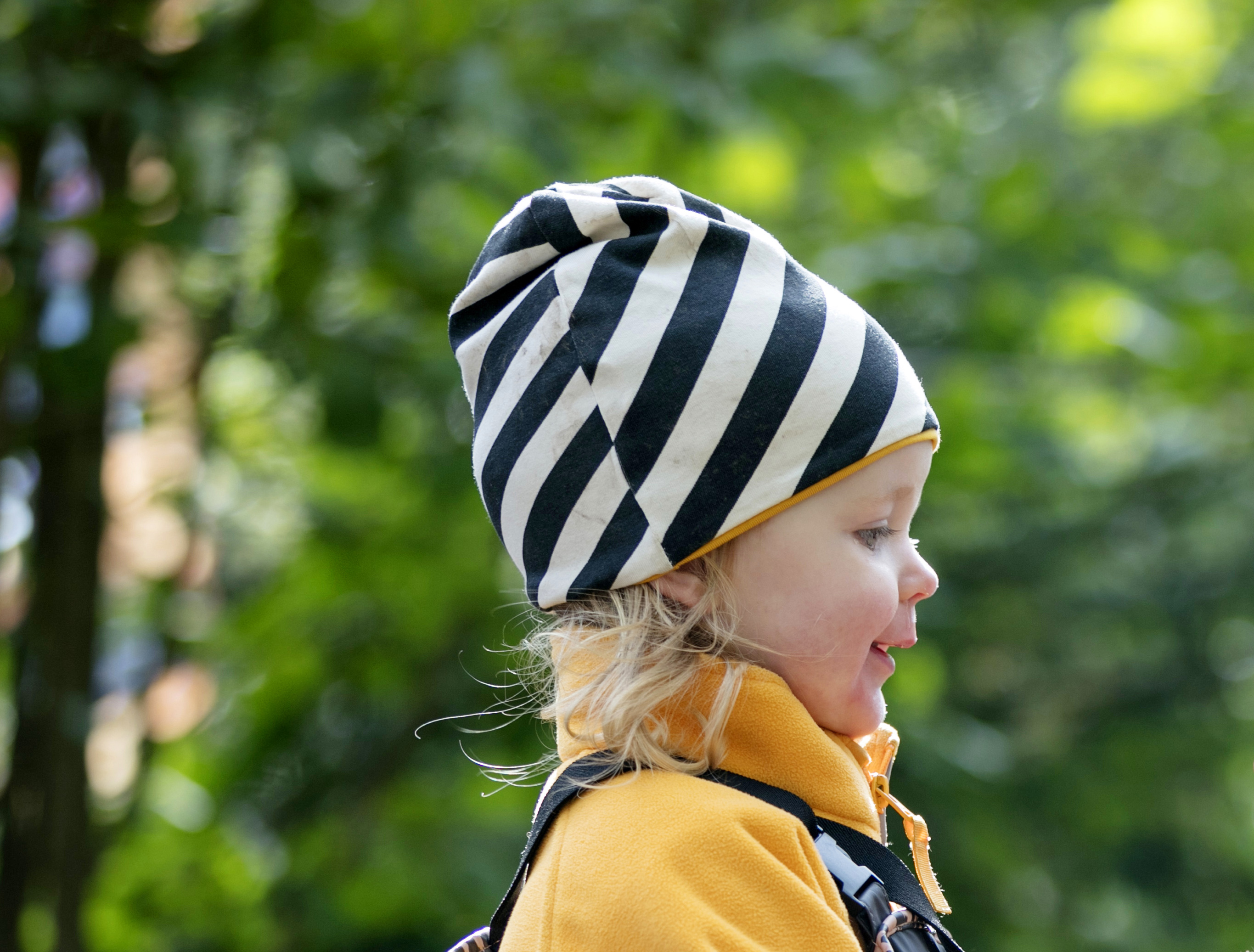 Liten blond flicka är ute i naturen. På sig har hon en randig, svart och vit mössa samt en gul fleecejacka. 