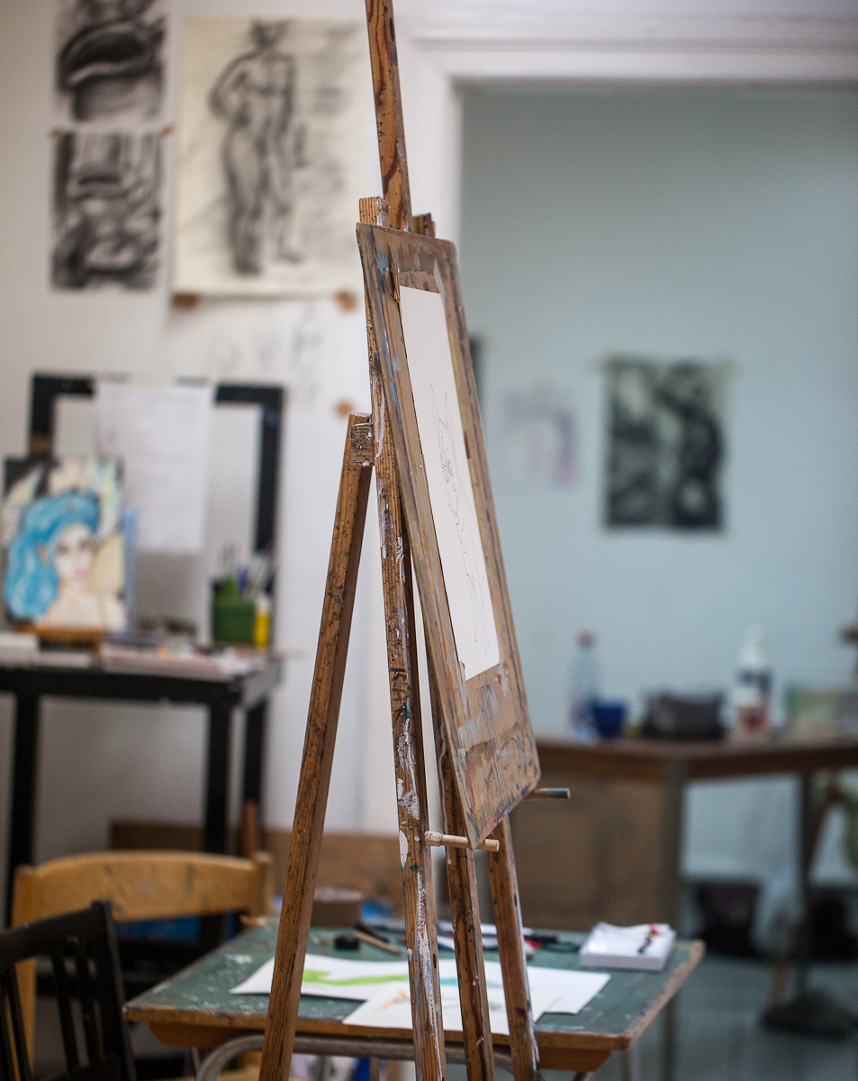 An empty easel in an art gallery. In the background you can see sketches and paintings hanging on the wall.