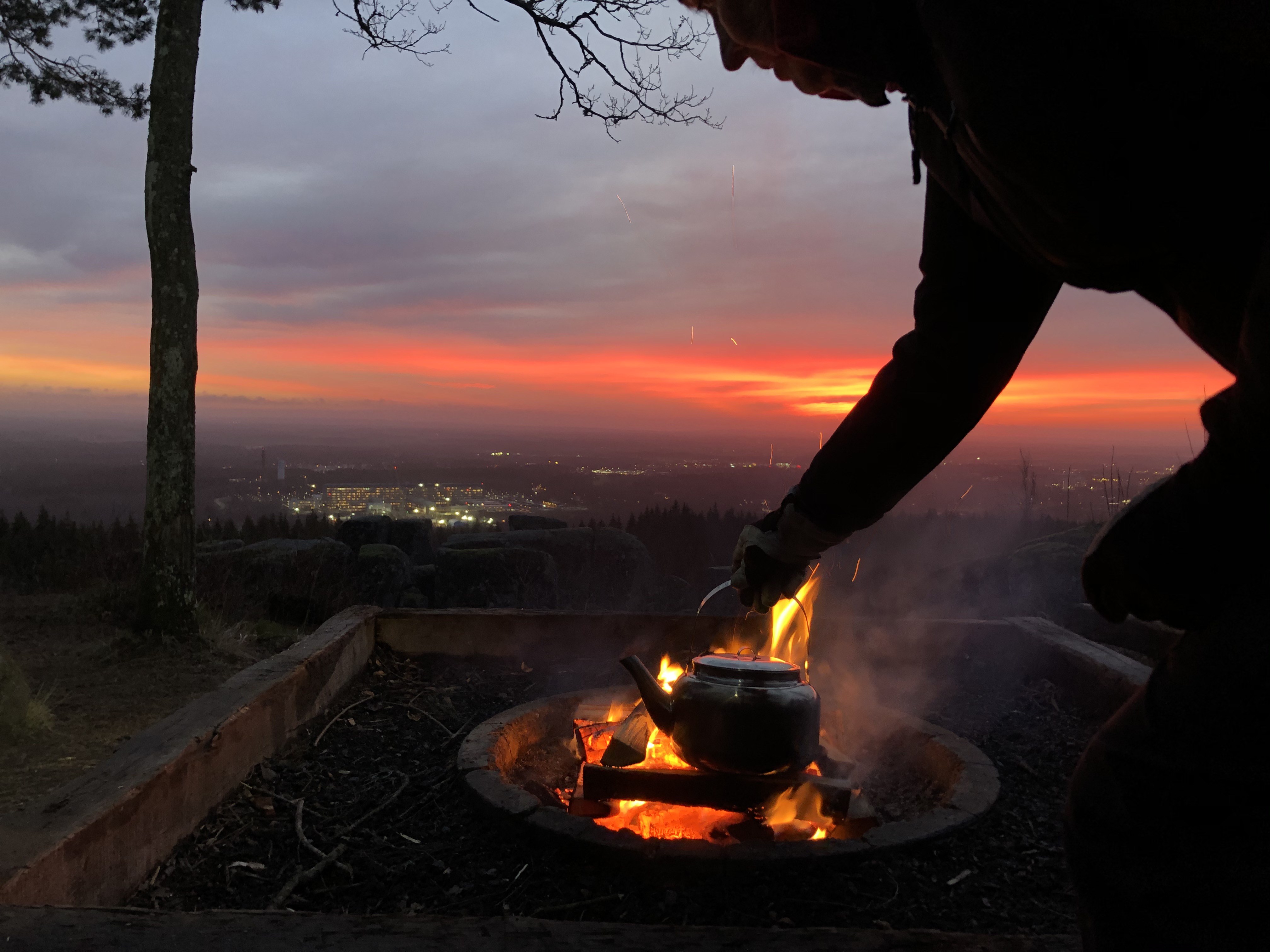 En kvinna fixar kaffet över öppen eld i soluppgången med utsikt över staden Skövde.