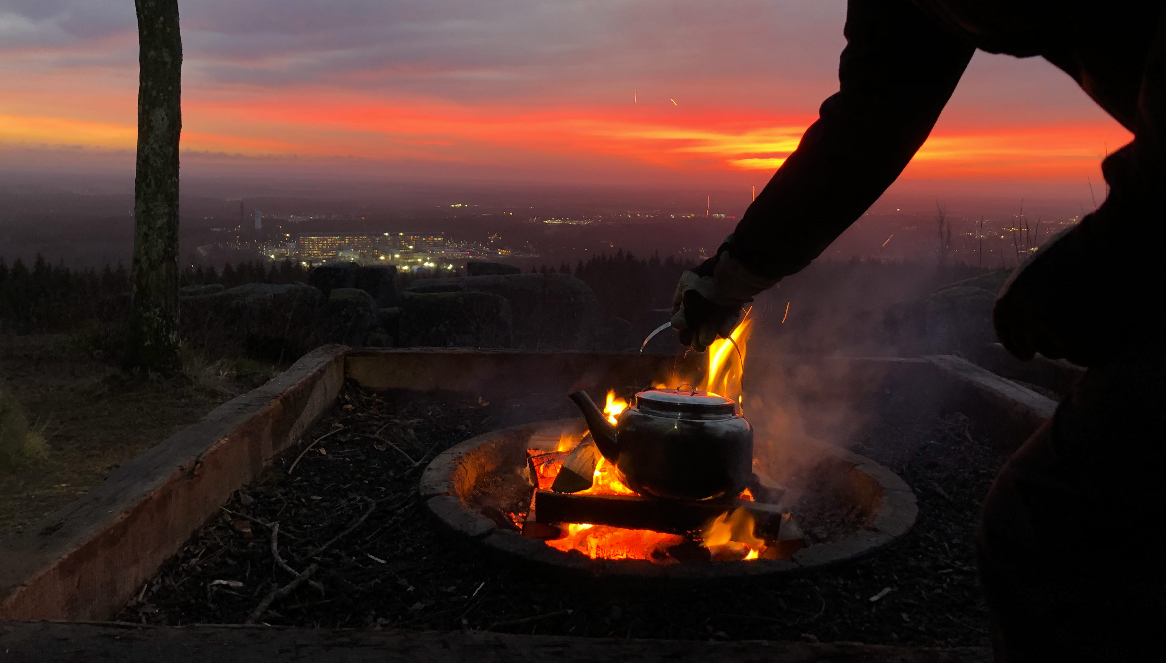 A woman fixes her coffee over an open fire at sunrise with a view of the city of Skövde.