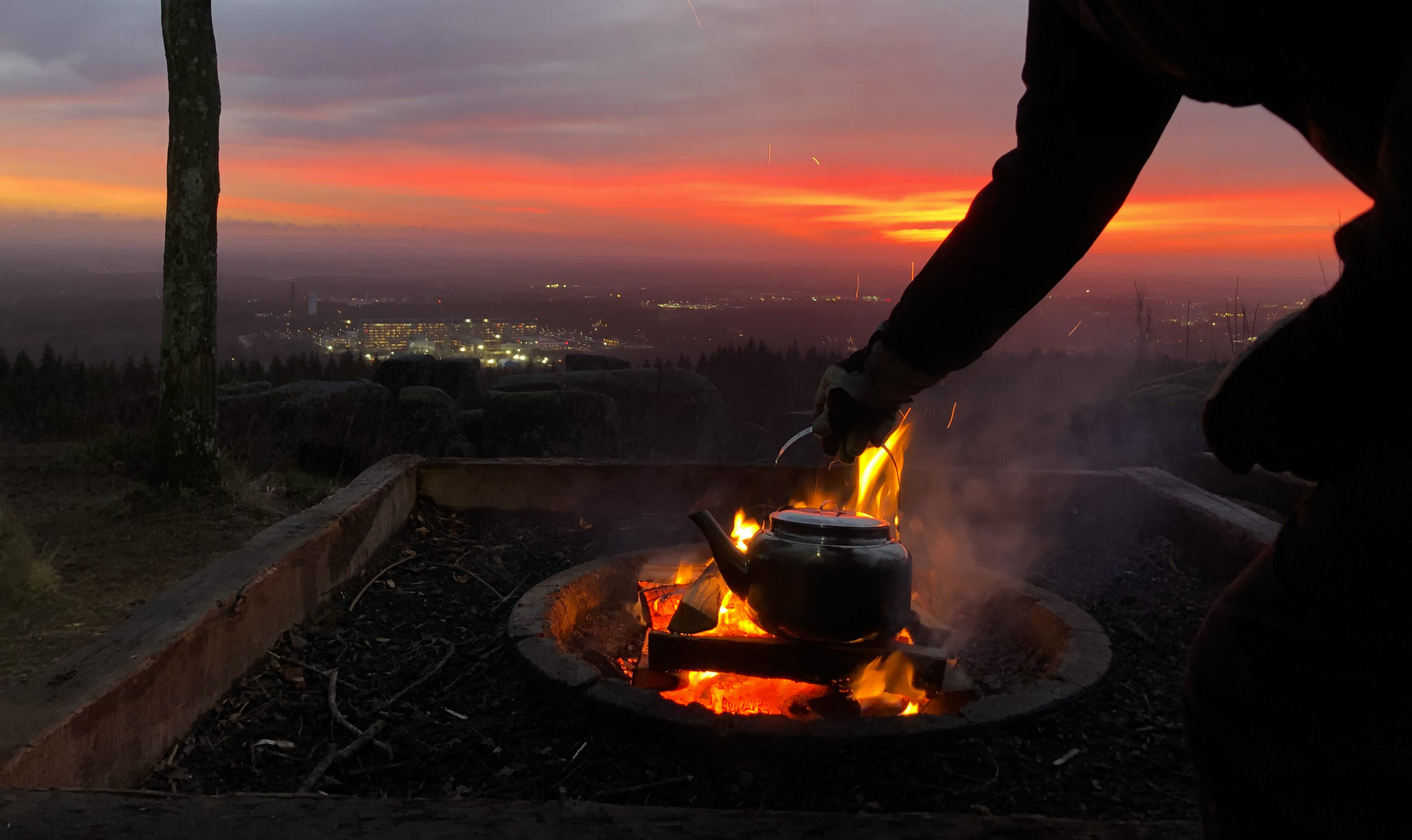 En kvinna fixar kaffet över öppen eld i soluppgången med utsikt över staden Skövde.