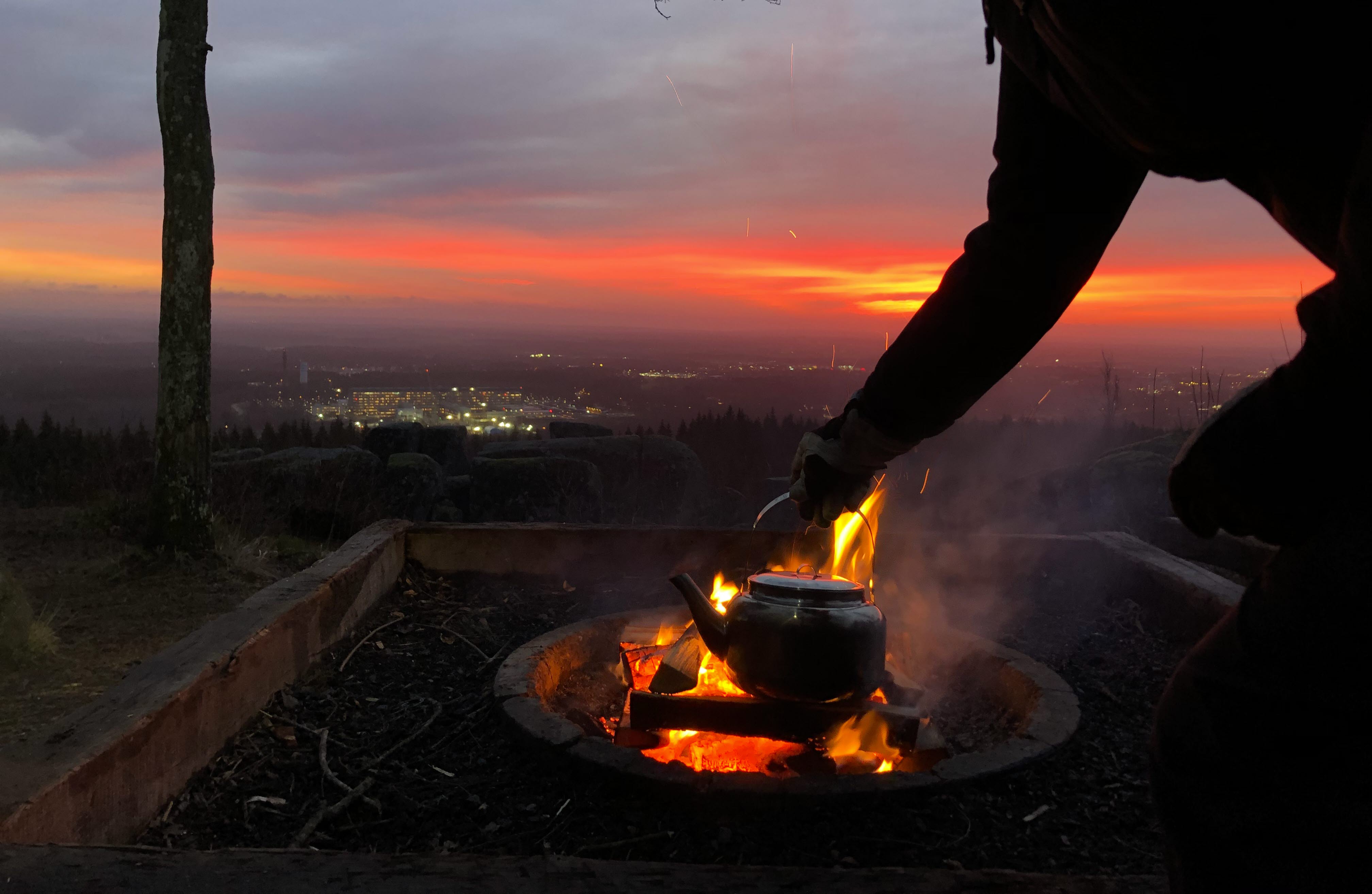 En kvinna fixar kaffet över öppen eld i soluppgången med utsikt över staden Skövde.
