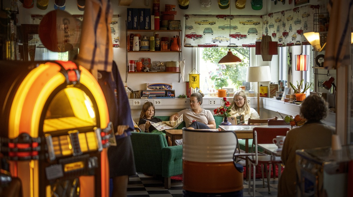 En mamma och hennes två tjejer i tonåren sitter i en grön soffa omgivna av retroprylar och en jukebox i ett café. 