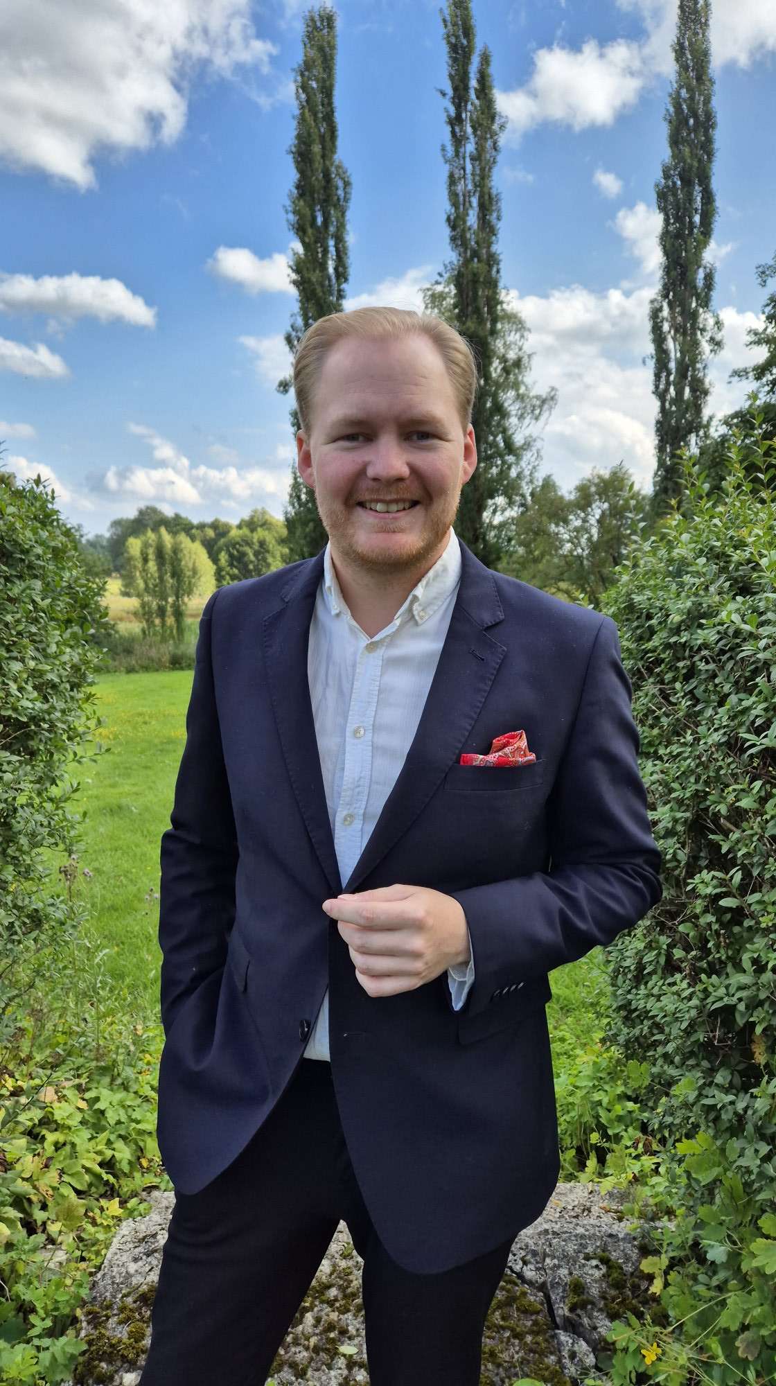 En man i blå kostym med vit skjorta i ett grönområde. 