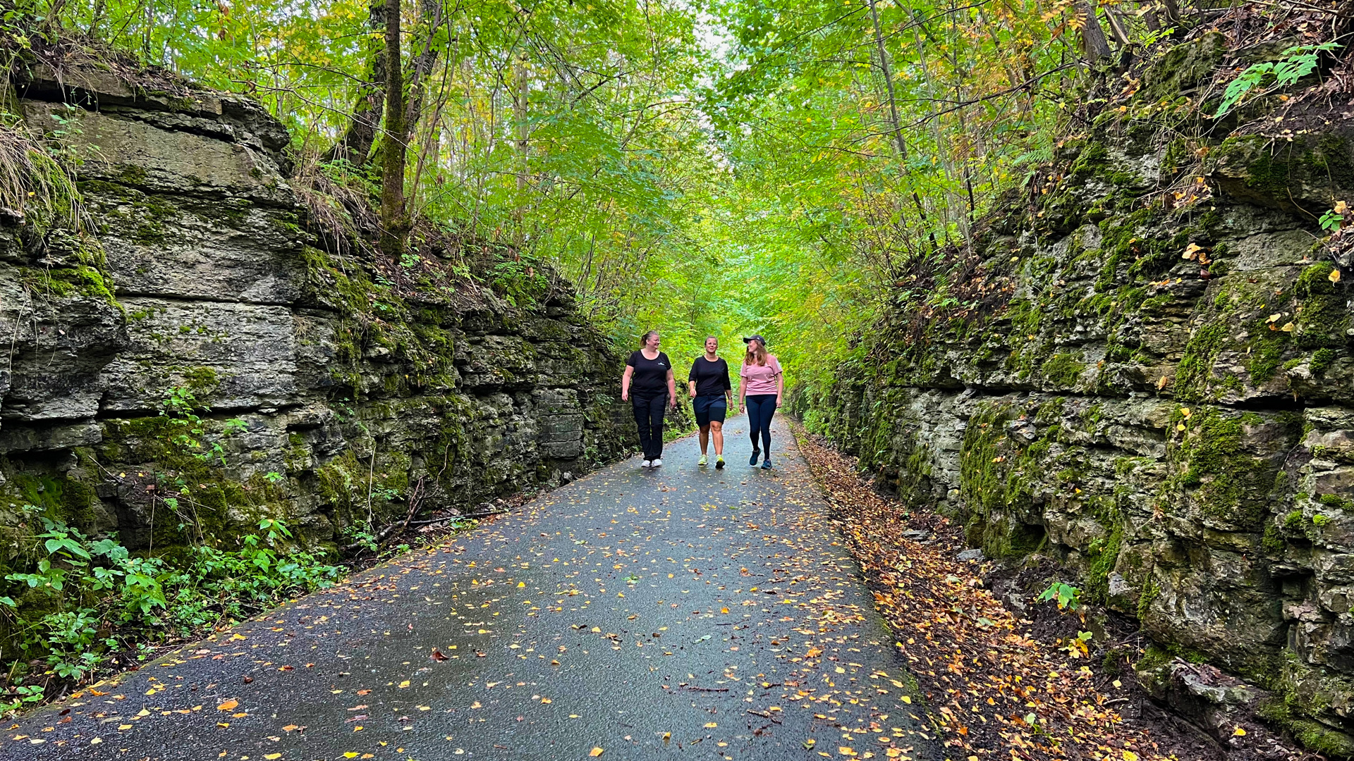 Tre kvinnor vandrar på en gammal banvall i ett höstfärgat landskap där bergets lager syns tydligt på bergsväggarna som omger vägen på banvallen. 