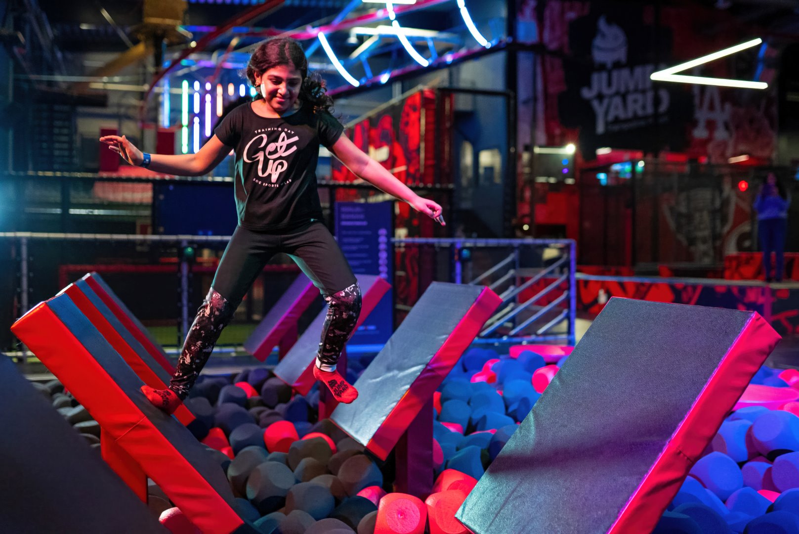 En flicka i svart hår och svarta kläder hoppar mellan hindren i en trampolinpark.
