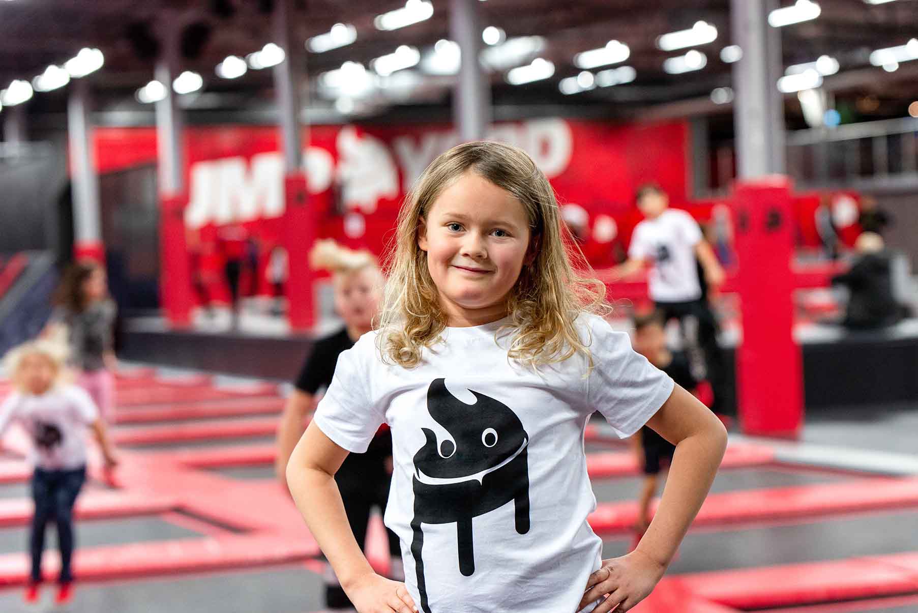 En flicka med blont hår och vit tröja tittar in i kameran. I bakgrunden ser man flera trampoliner från en trampolinpark breda ut sig. 