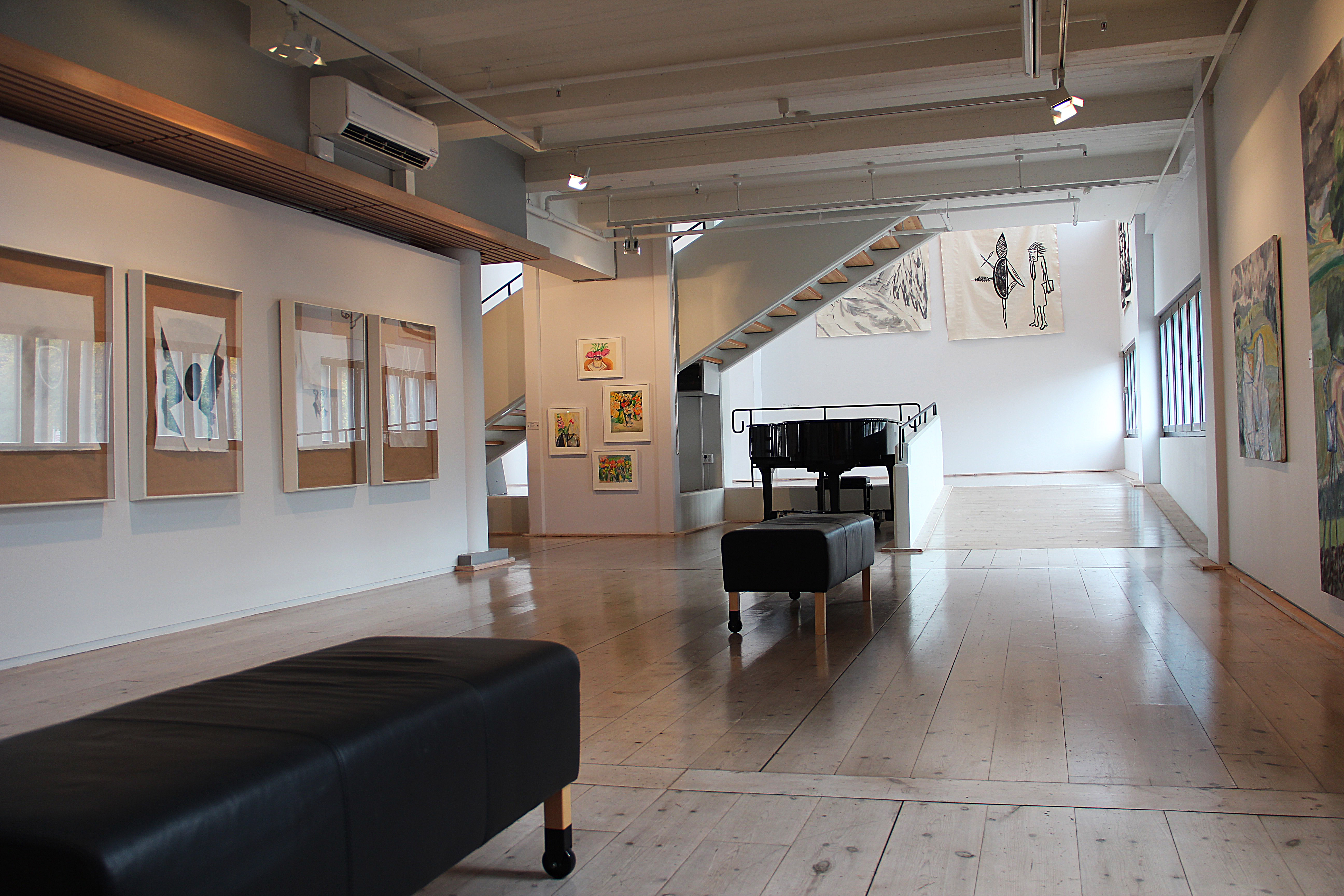 Stor vitmålad lokal. På väggarna sitter ramar med konst i. Längre in i bild skymtar ett svart piano. 