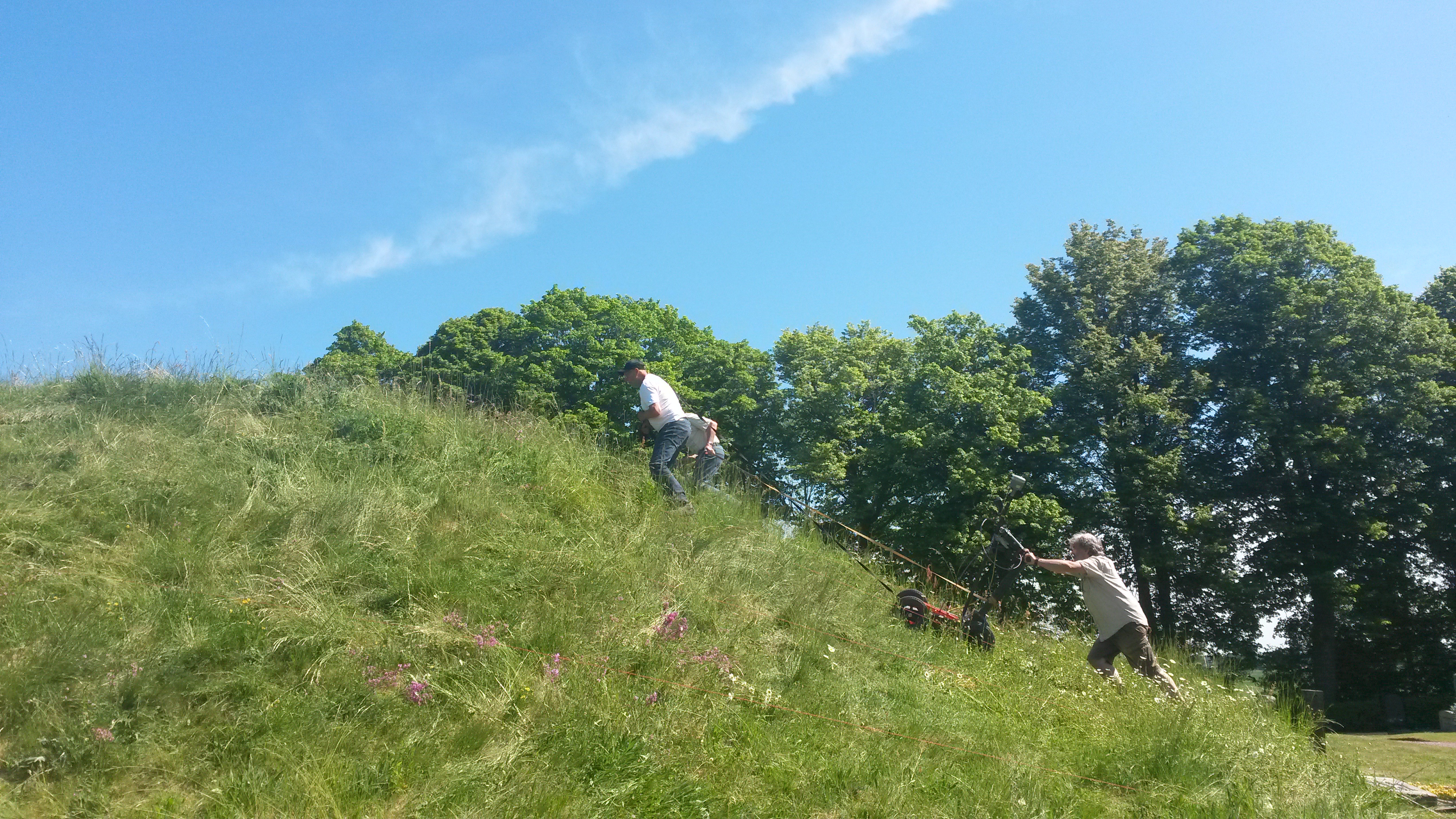 Tre personer släpar ett undersökningsinstrument upp för en gravhög som de utför en markradarskanning av. 