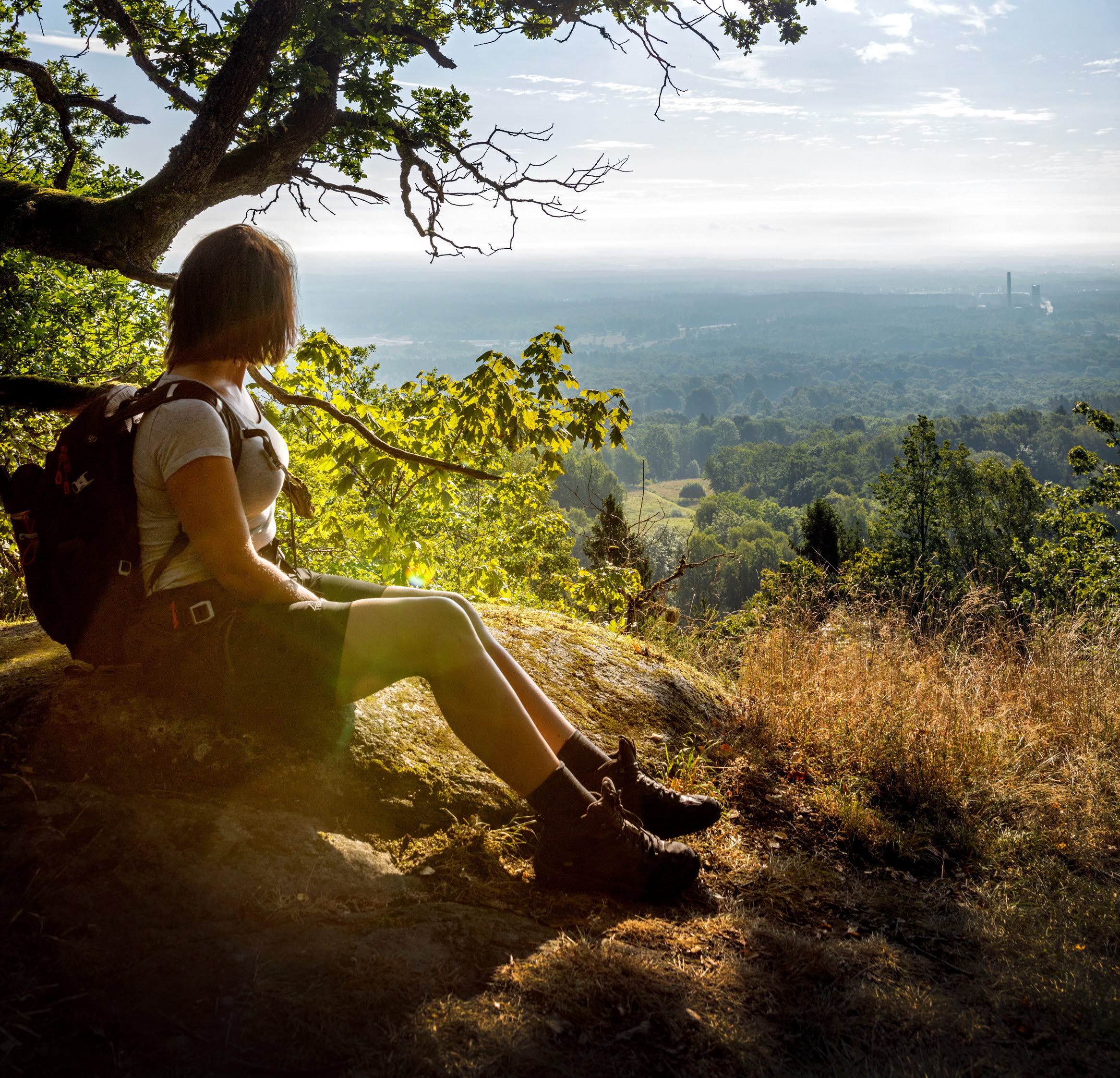 Kvinna sitter på sten i shorts och t-shirt med ryggsäck på ryggen och tittar ut mot utsikten till hennes vänster. Solen glittrar genom i trädets grenar snett bakom henne. 