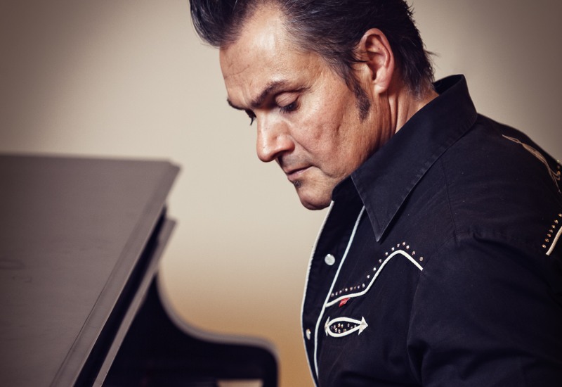 En man med polisonger och bakåtkammat hår vid ett piano.