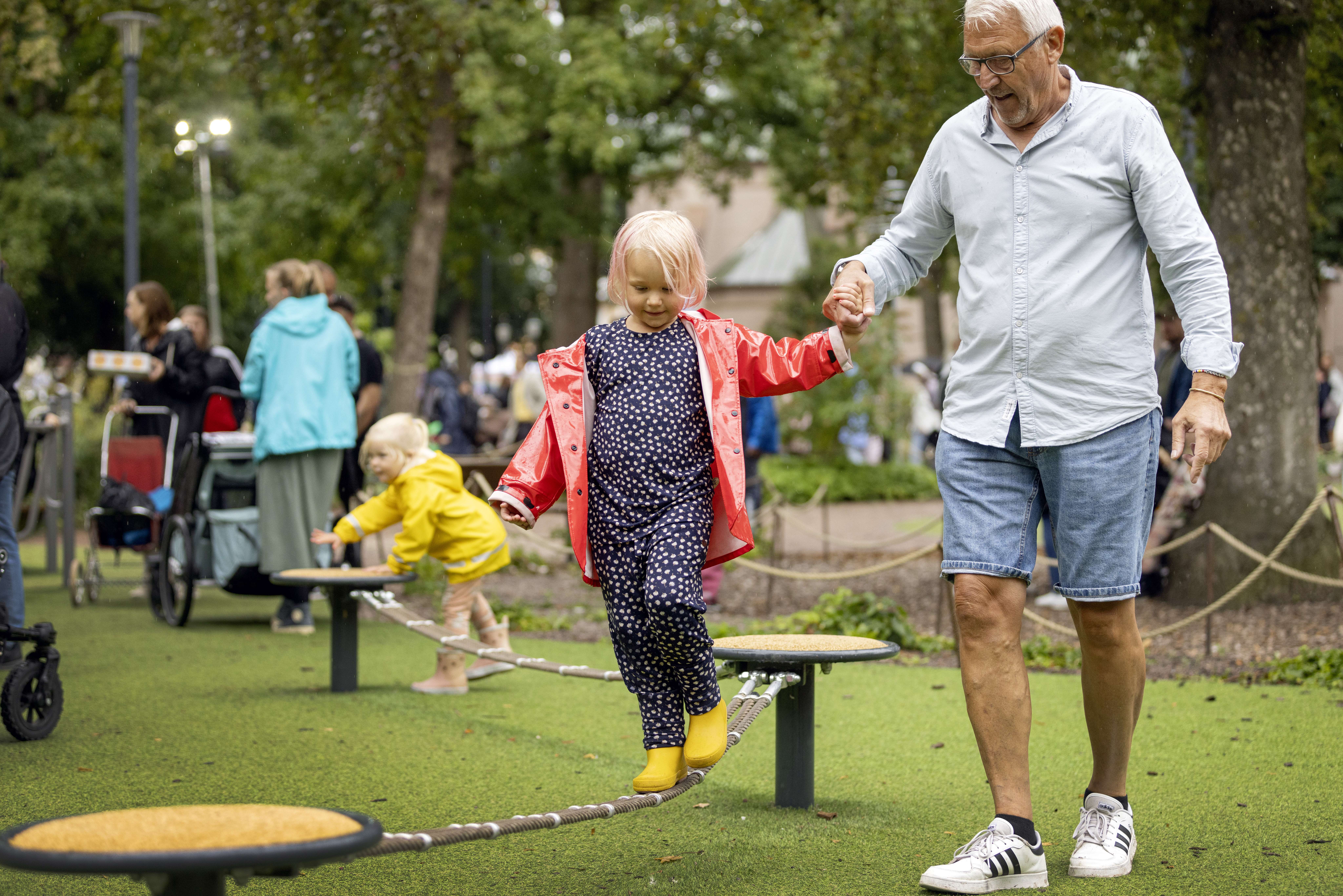 En liten flicka går balansgång på en lekplats med en äldre man som hjälper henne att hålla balansen.