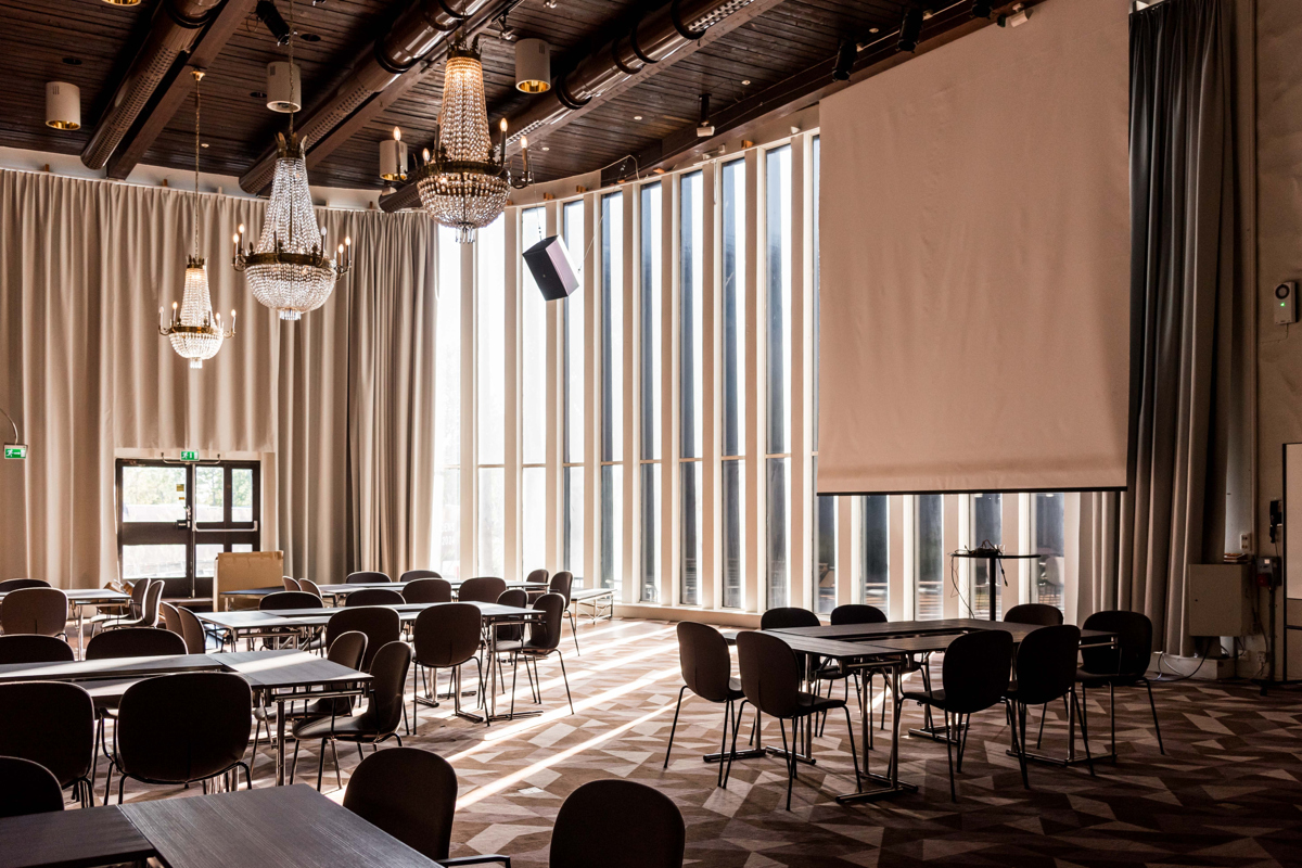 En bild som visar en konferenslokal med höga fönster, stolar och bord i grupper och en projektorduk. 