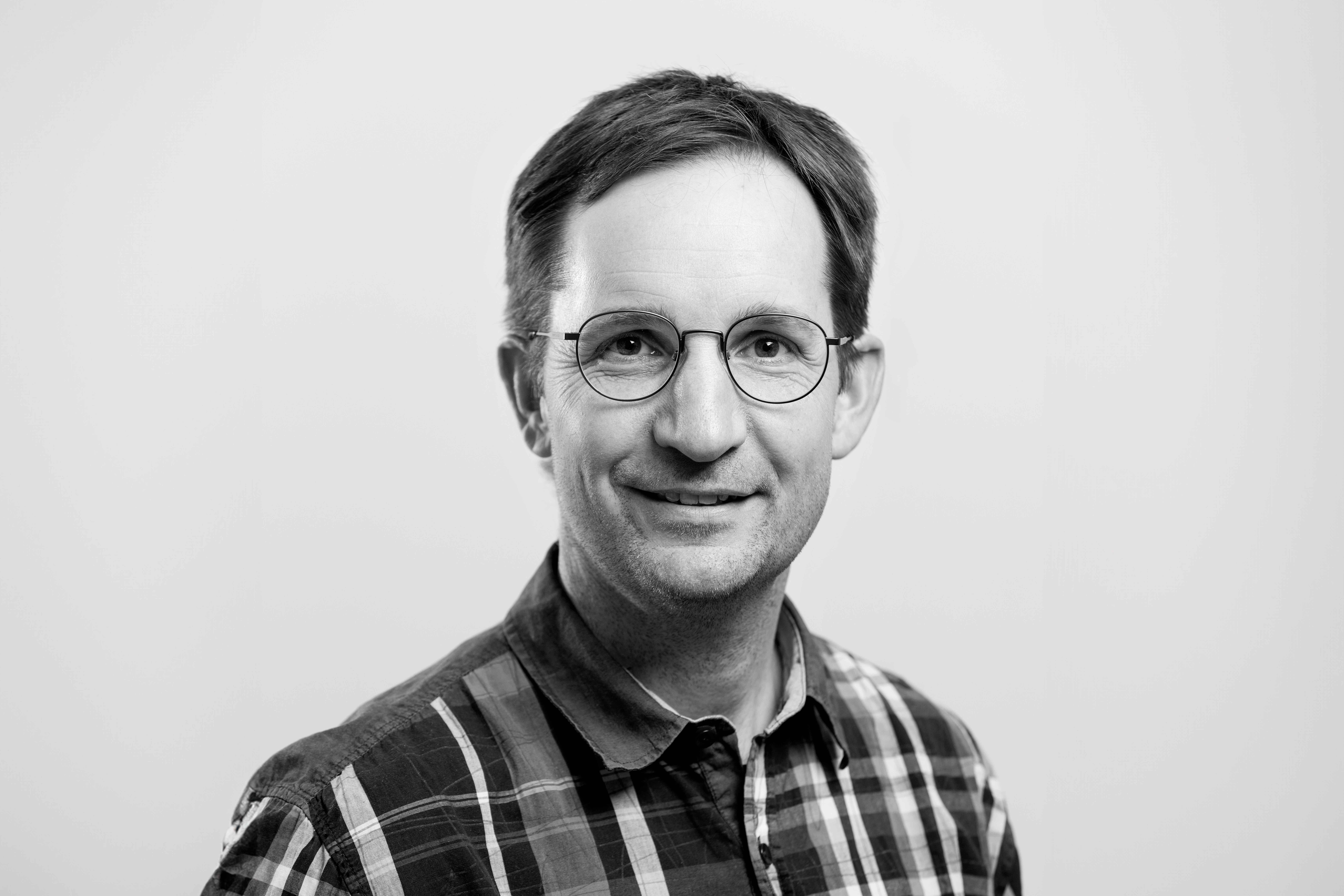 Ett svartvitt porträtt av en man med mörkt hår, glasögon och rutig skjorta. 