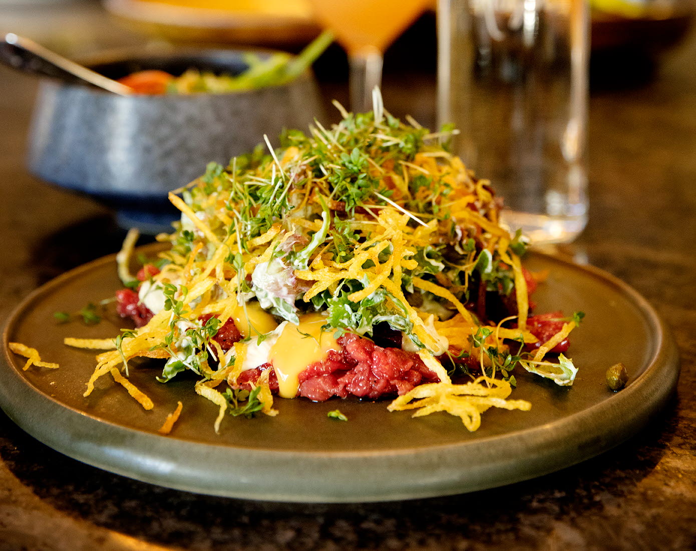 En tallrik med råbiff och tillbehör fint upplagt på råbiffen. I bakgrunden syns en drink, en vattenflaska och kocken. 