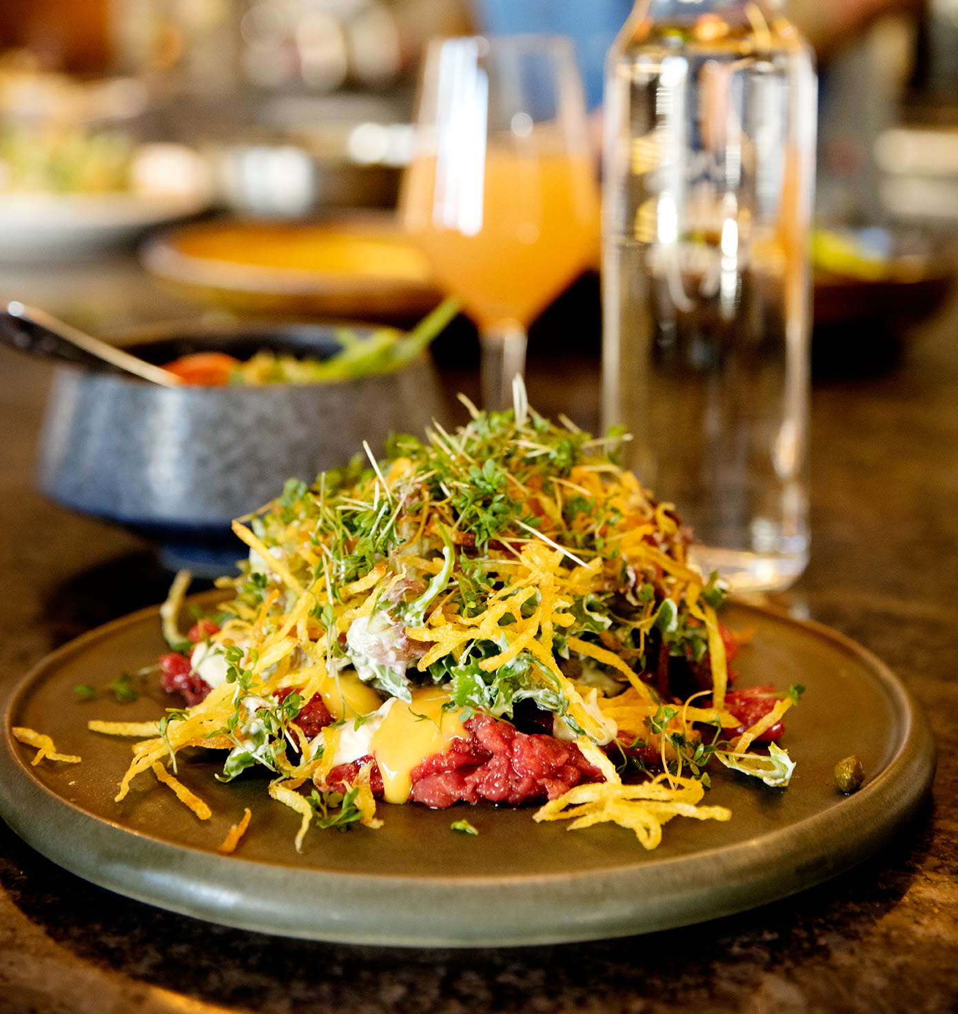 En tallrik med råbiff och tillbehör fint upplagt på råbiffen. I bakgrunden syns en drink, en vattenflaska och kocken. 