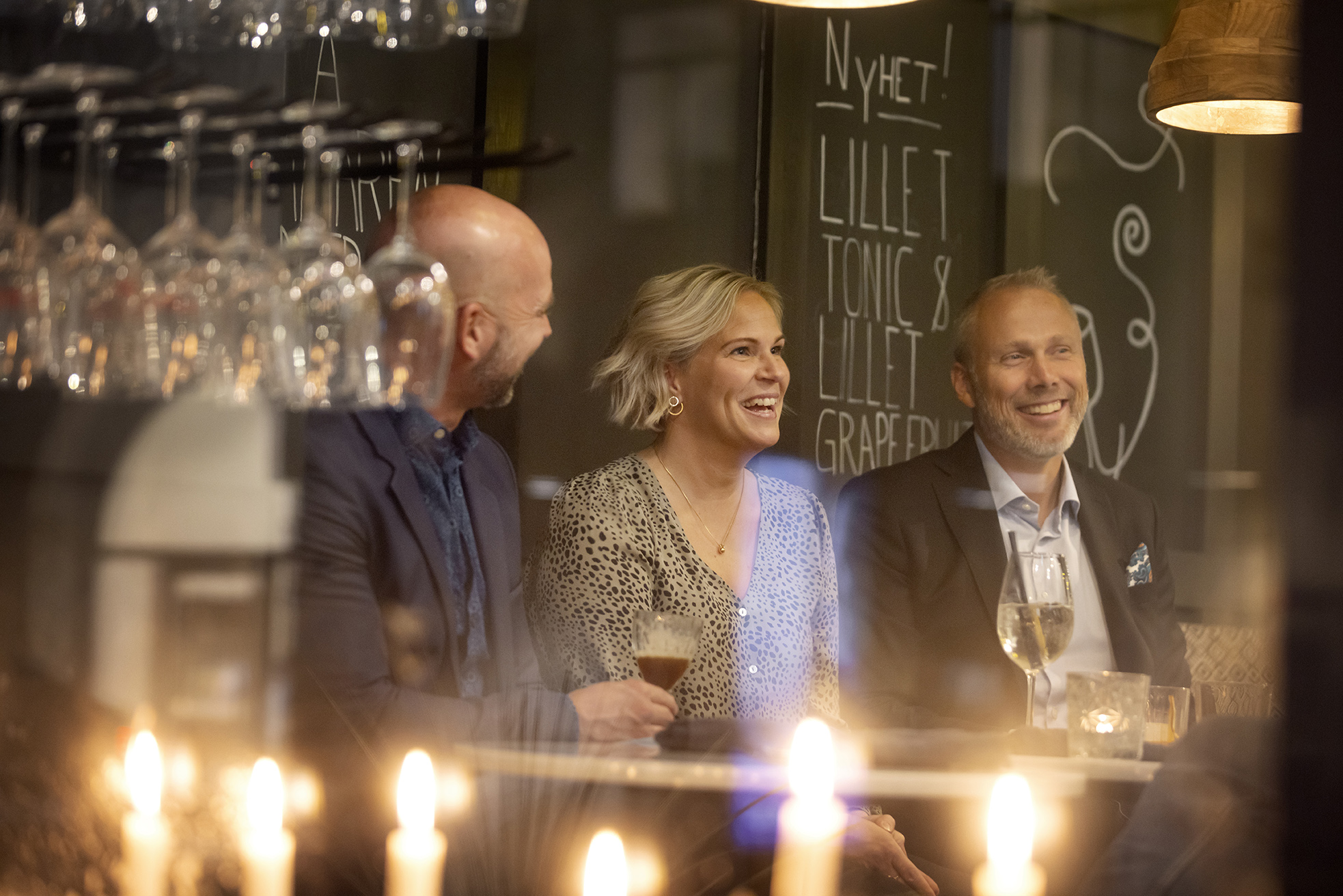 Tre personer i en bar. Glas och tända ljus i förgrunden. 