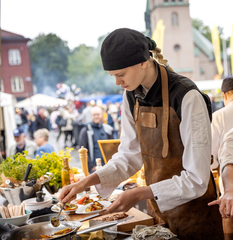 Tre tjejer med kockförkläde som lagar mat på ett torg med kyrka i bakgrunden.