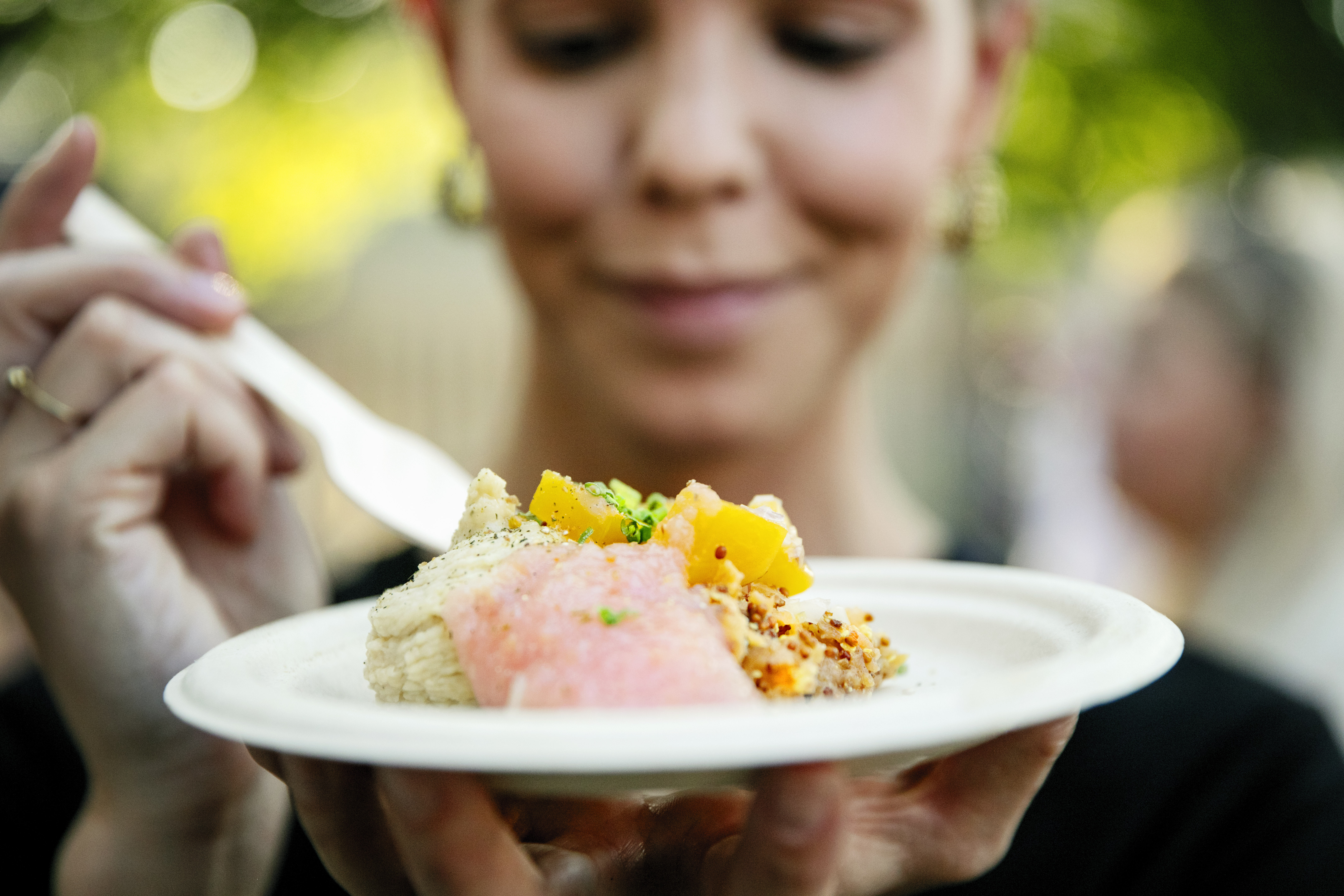 I förgrunden ser man en tallrik med mat och i bakgrunden en tjej som håller man ett bestick för att äta.