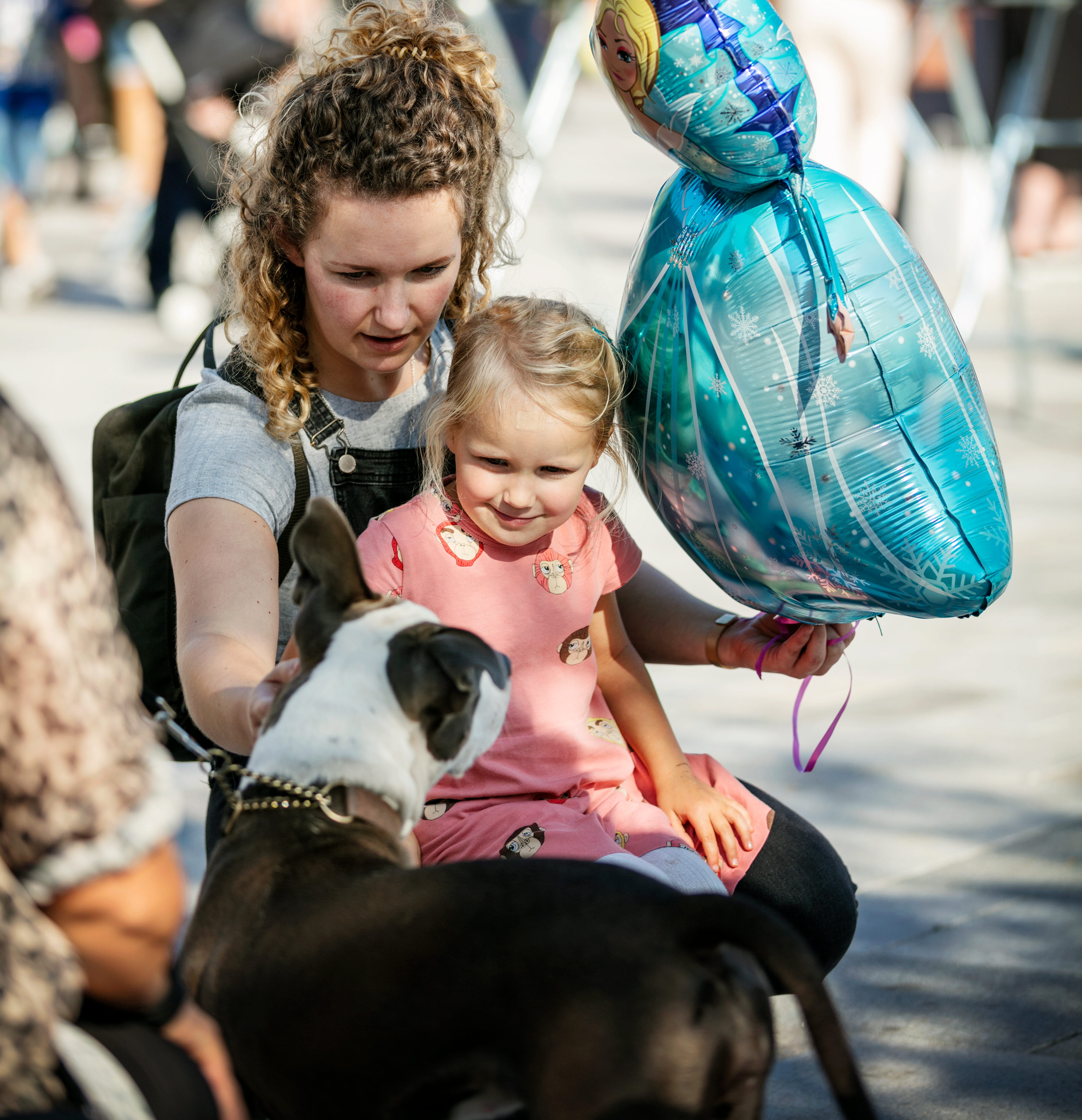 En flicka sitter i en kvinnas knä och klappar en brun-vit-fläckig hund samtdigt som kvinnan håller en blå ballong. 