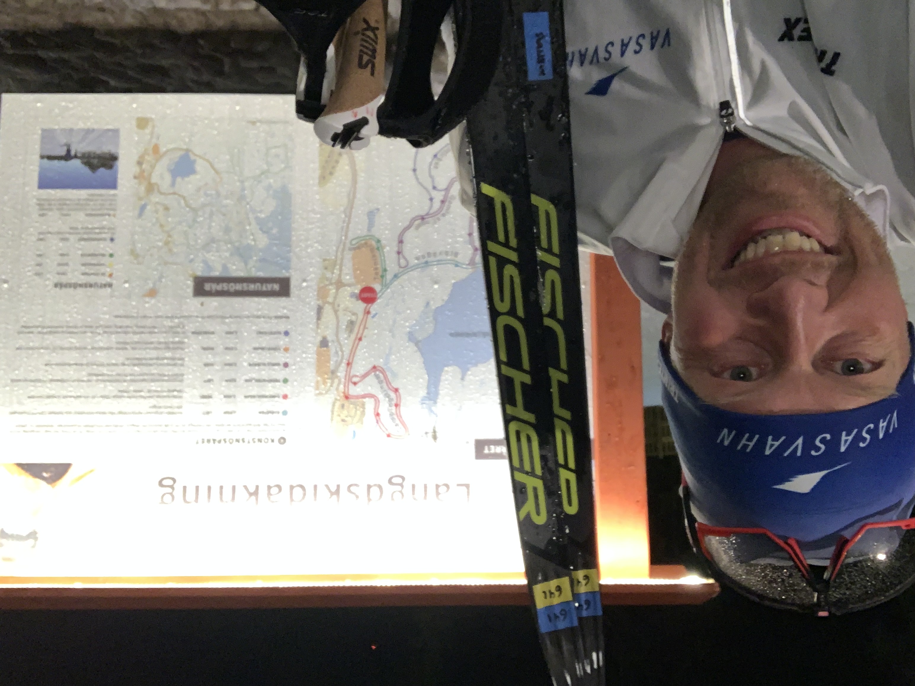 Skidåkaren Mattias Svahn i mössa, skidglasögon och med skidorna i handen. Bakom honom syns en karta över längdskidspåren. 