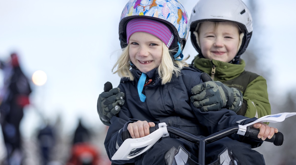 A girl with a colorful hat and helmet rides a snowracer with a boy sitting behind her wearing a green jacket and white helmet.