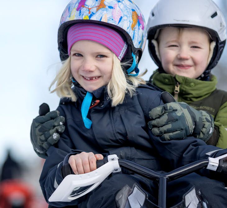 En flicka med färgglad mössa och hjälm åker snowracer tillsammans med en pojke som sitter bakom henne iklädd grön jacka och vit hjälm.