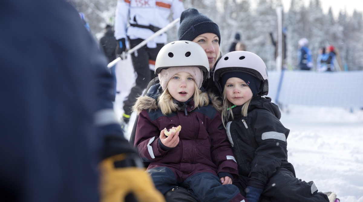 Två flickor sitter i vuxens knä utomhus på vintern iklädda overall och hjälm och äter korv med bröd.