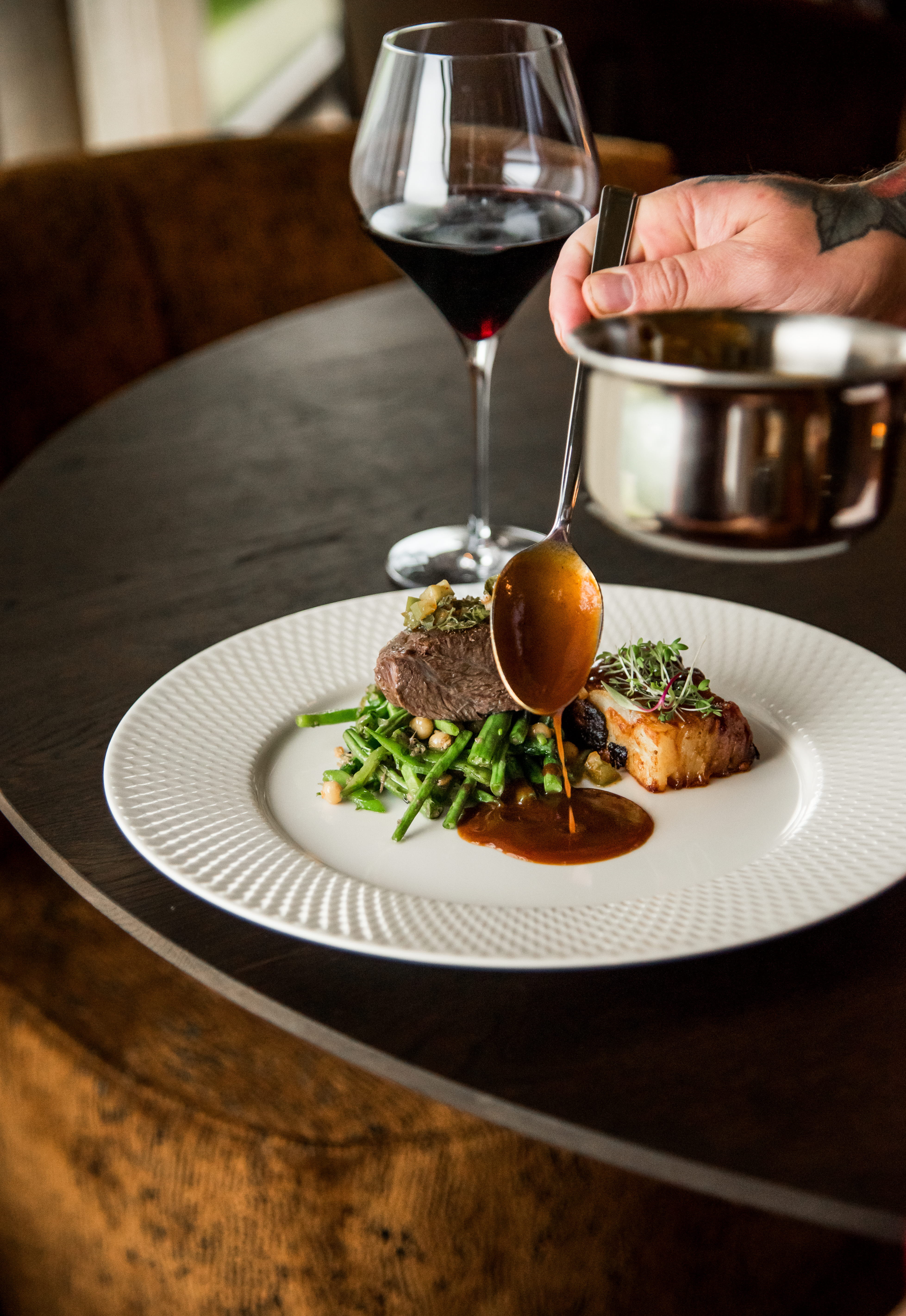 En hand hållandes i en sked häller upp sås på en tallrik med gröna bönor och kött. Bakom tallriken står en glas med vin och intill syns en kastrull. 
